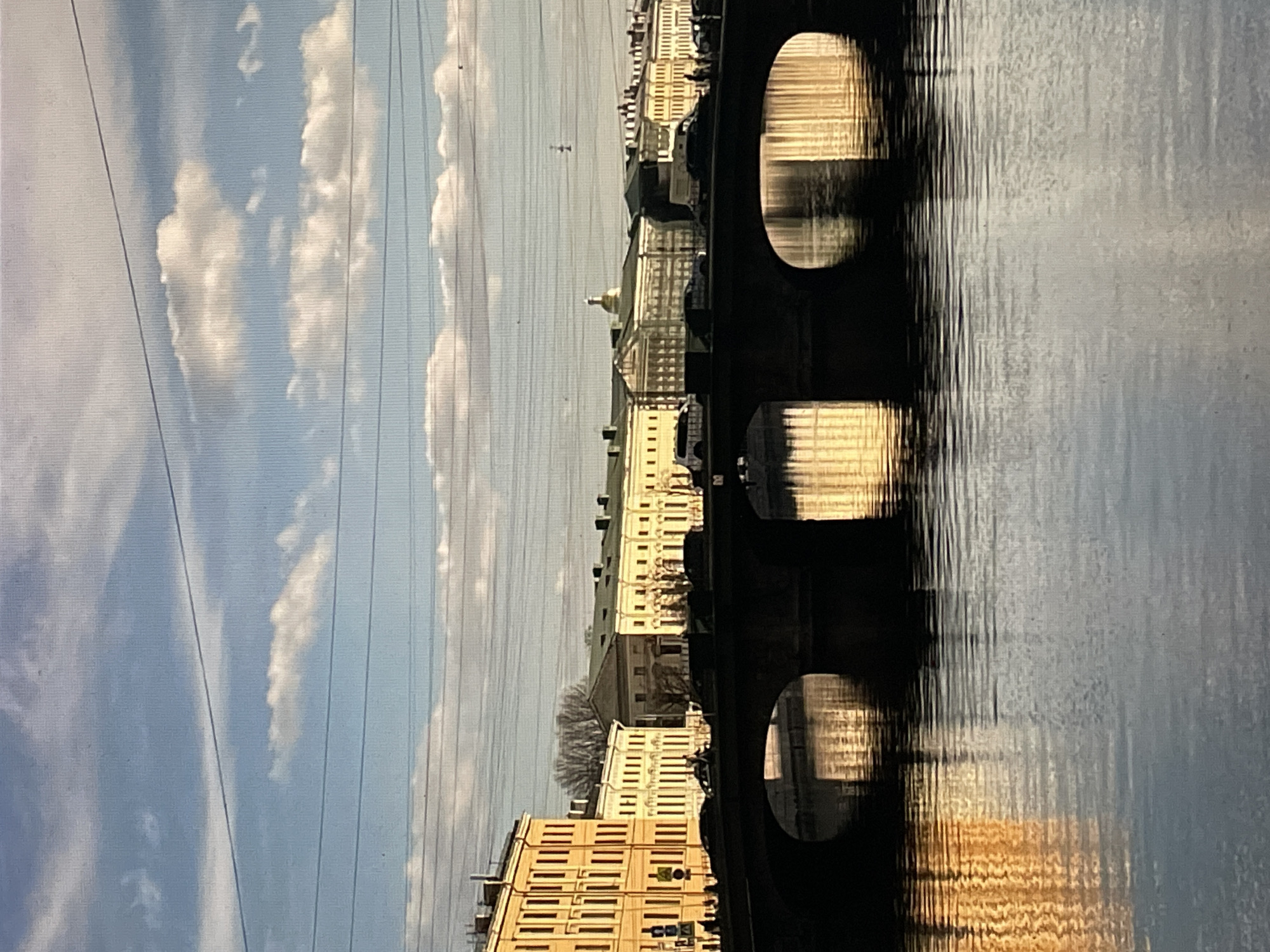 The image displays an urban landscape in Saint Petersburg, Russia, capturing a view along the Moyka River looking east-southeast from or near the Dvukharkovy Most (Two-Arch Bridge).
In the foreground, the Moyka River's dark, calm water reflects the sky and sunlit building facades, with surface ripples causing minor distortion. The Dvukharkovy Most, a stone two-arch bridge, spans the river as a dark silhouette, its two arches creating shadowed openings beneath.
On the bridge deck, several dark-colored passenger cars and vans are visible, some stationary, others in apparent motion. Indistinct human figures are present on the bridge's sidewalks and along the river embankments.
Multi-story historical buildings line both the left (south) and right (north) embankments. Those on the left are prominently illuminated in a warm yellow/ochre hue, characterized by numerous vertically aligned windows. Further along the right embankment, similar structures extend into the distance, including one building featuring a classical pediment and columnar facade.
In the background, the large, golden dome of St. Isaac's Cathedral rises above the distant rooftops, catching direct sunlight against a light blue sky. The sky contains scattered, horizontally elongated white cumulus clouds. Multiple thin, dark horizontal wires, likely utility lines, traverse the upper portion of the image.
The scene is illuminated by strong, low-angle sunlight, producing deep shadows under the bridge and brightly highlighting the building facades and the cathedral dome.