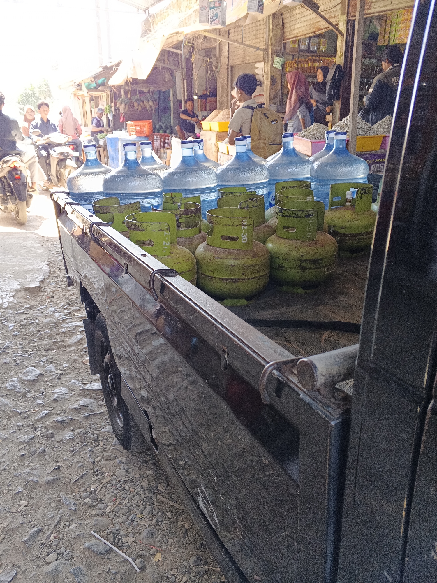 A black pickup truck is parked on a dirt and gravel road in a marketplace area. The bed of the truck is loaded with large blue water jugs and several green propane tanks. In the background, there are stalls and shops displaying various goods, including what appear to be dried fish or shrimp, vegetables, and bottled products. Several people are visible, some walking, some on motorcycles, and some tending to the stalls. The scene suggests a busy market day in Warungkiara, Indonesia, with the truck likely delivering or collecting supplies. The lighting indicates it is daytime, with sunlight filtering through overhead awnings.