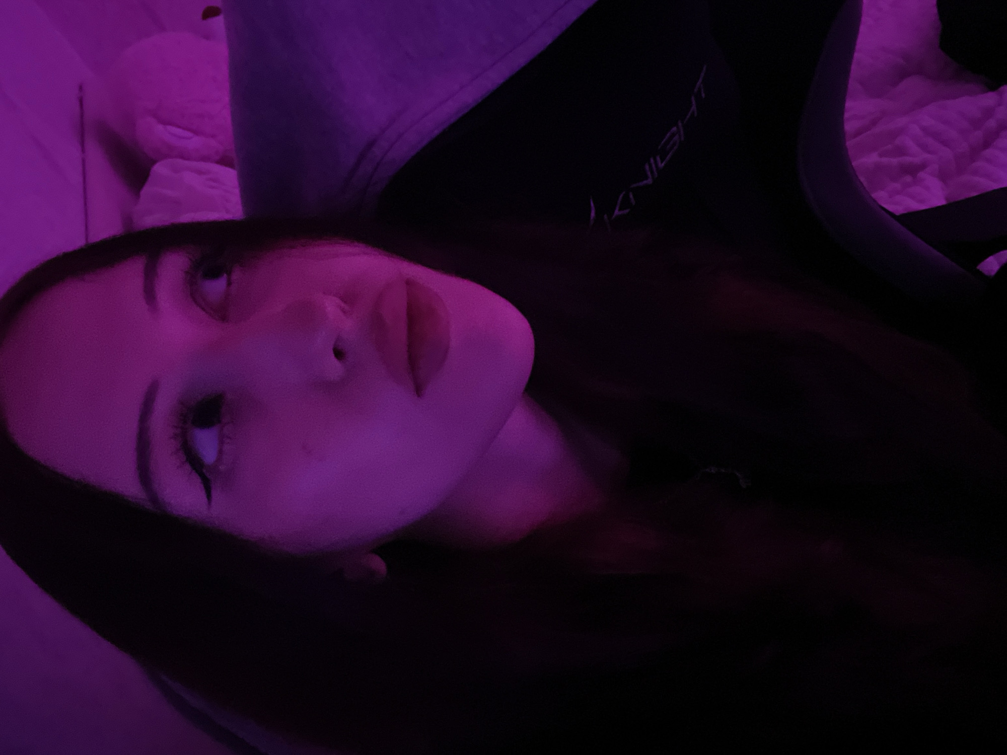 The scene is located in Votkinsk, Russia. A female subject, approximately from the chest up, is captured in an indoor setting under dominant magenta and purple artificial lighting. She has long, dark hair, winged eyeliner, and accentuated eyelashes. Her lips are full and slightly parted. Her gaze is directed upwards and to the right of the frame, fixated on an unseen point. She wears a dark garment, possibly a hoodie, upon which the embroidered or printed word "KNIGHT" is visible in a light color on the left side of the frame, angled downwards. To the right of the subject, in the background, multiple soft, light-colored objects consistent with pillows or plush toys are visible, also bathed in the pervasive purple light. Below her right arm (viewer's left), a textured white material, consistent with bedding, is present. The overall lighting scheme creates strong contrasts and an atmospheric, low-light environment.