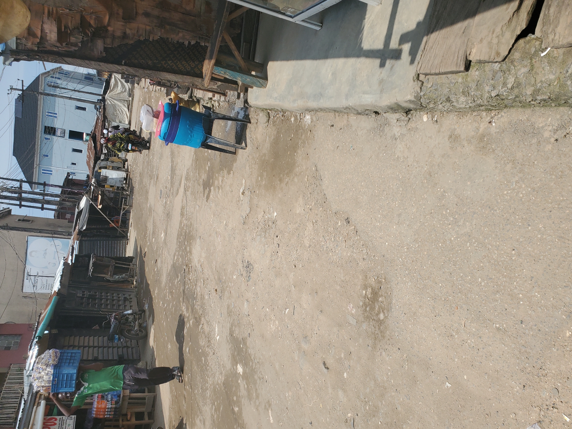 The image depicts a street scene in Shangisha, Nigeria, characterized by a dusty, unpaved road with scattered debris and puddles of water. In the foreground, a man in a green shirt and black pants carries a blue crate filled with bottled drinks on his head. He is walking away from the viewer towards the left side of the frame. To his left, a small market stall with a corrugated metal roof is visible, with some beverages stacked on a table. A motorcycle is parked in front of the stall.
Further down the street, two motorcycles are approaching, carrying riders. One rider is wearing a colorful patterned top, and the other is wearing a white helmet. Behind the motorcycles, a pale blue multi-story building with solar panels on the roof dominates the background. The buildings lining the street appear to be a mix of market stalls and residential structures, some with corrugated metal roofing. A large blue water cooler sits on a stand on the right side of the road, in front of a brick wall and a wooden structure. The overall impression is one of a busy, informal commercial area in an urban setting.