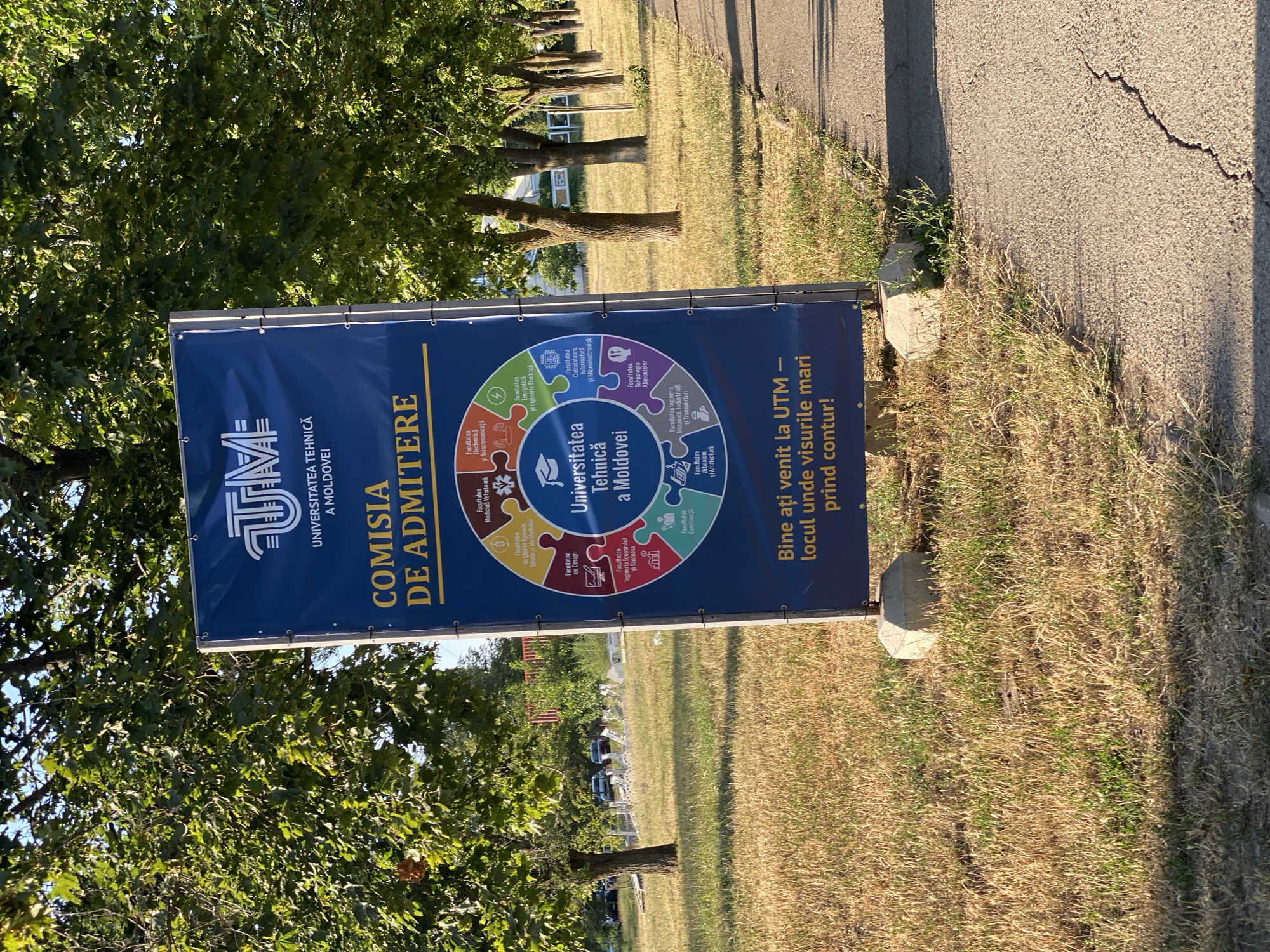 The image shows a large blue banner sign standing on a concrete base in an outdoor setting. The banner features the logo of UTM (Universitatea Tehnică a Moldovei - Technical University of Moldova) and text in Romanian. The text on the banner reads "COMISIA DE ADMITERE" (Admissions Committee) and lists various faculties of the university with their respective icons, arranged in a circular diagram around the university's name and location, "Universitatea Tehnică a Moldovei". Below this section, there is a slogan: "Bine ați venit la UTM — locul unde visurile mari prind contur!" (Welcome to UTM - the place where big dreams take shape!).
The sign is situated on a patch of dry, yellowed grass, with a cracked asphalt path running alongside it. In the background, there are several trees with green foliage, and a row of more trees can be seen further in the distance. The sky is visible above the trees, suggesting it is daytime. Some cars are parked in the background, near what appears to be a building or a fenced area. The overall impression is an entrance or information point for the technical university, likely in Chișinău, Moldova, given the university's name. There are no people visible in the frame.