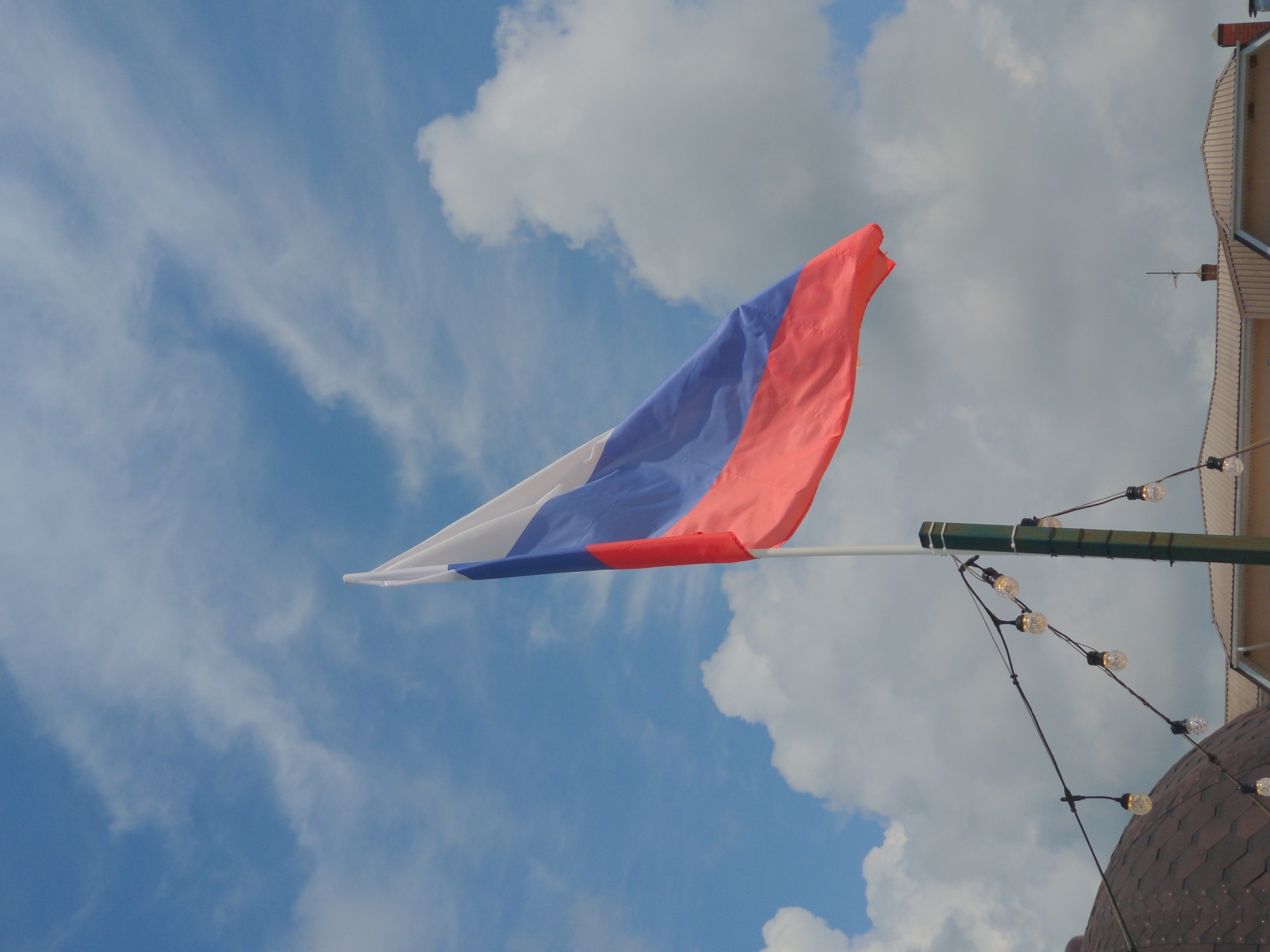 A Russian flag, white, blue, and red, is captured mid-air, waving against a cloudy blue sky. The flag is attached to a pole, which is positioned to the right of the frame. In the foreground, a strand of fairy lights dangles from wires strung across the scene. Below the lights, the dark, domed roof of a building is visible on the left side of the image, and further to the right, the light-colored facade of another building with a tiled roof can be seen. The overall scene is set outdoors, under a daytime sky.