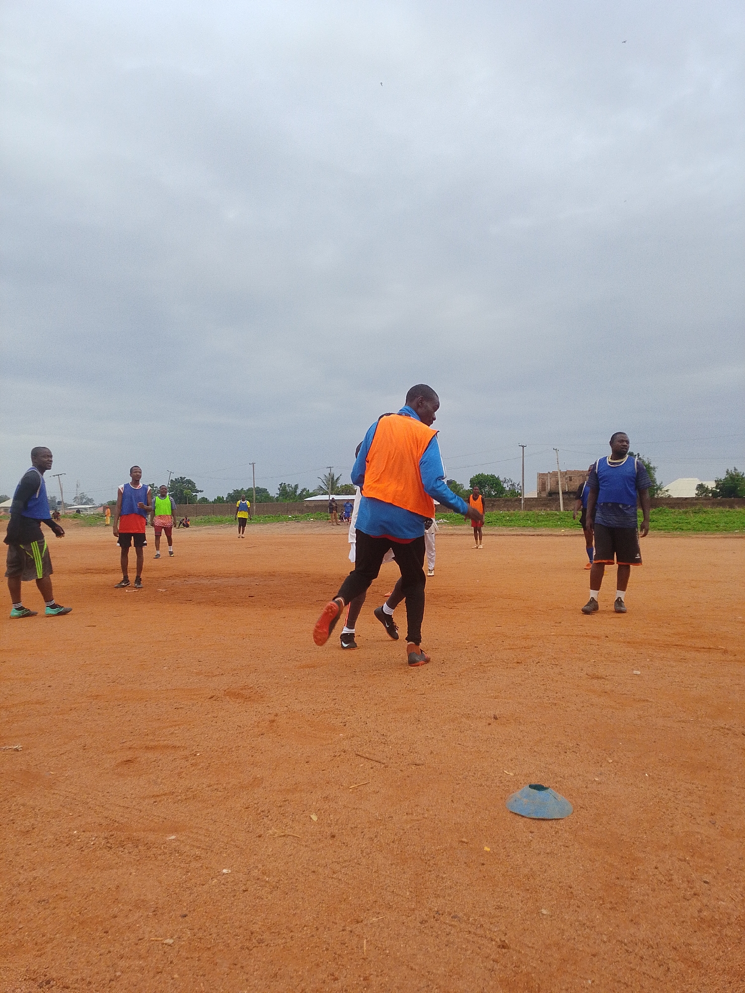 On an outdoor, brown dirt field in Bukuru, Nigeria, a group of men is participating in a soccer training session. The sky is overcast and gray. There are seven visible men in athletic attire and training vests of various colors (orange, blue, green, yellow), scattered across the field. One man wearing an orange vest is in mid-stride, facing away from the viewer. Another man stands stationary, wearing a blue vest. Other men are standing or walking in the background. In the foreground, a single blue cone sits on the dirt. The edge of a vegetated area, small structures, and utility poles are visible in the distance.