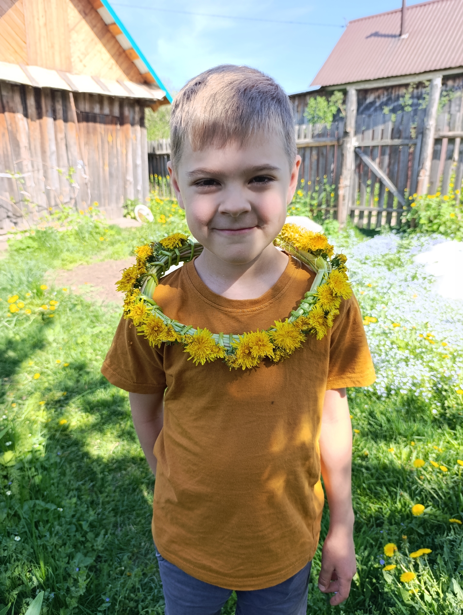 A boy stands in a grassy yard, wearing a dandelion wreath around his neck and a mustard-colored t-shirt. He has short, light-brown hair and a slight smile. Behind him, there is a wooden building with a blue-painted roof and a weathered wooden fence. The yard is filled with dandelions and small white flowers. The location is the city of Chaikovsky, Russia.