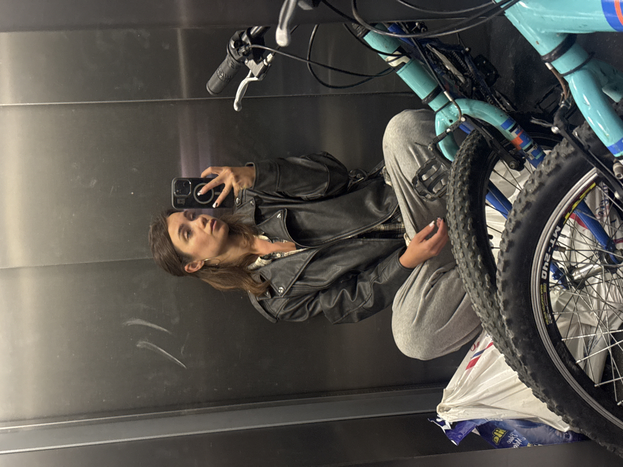 A woman in a black leather jacket and grey sweatpants is taking a selfie in a mirrored elevator, which appears to be located in Budva, Montenegro, based on the presence of a blue and white plastic bag with Cyrillic script possibly indicating a local store or brand. A turquoise bicycle is also inside the elevator, positioned between the woman and the mirror. The background is a plain, dark gray surface of the elevator walls. The woman is holding a black smartphone.