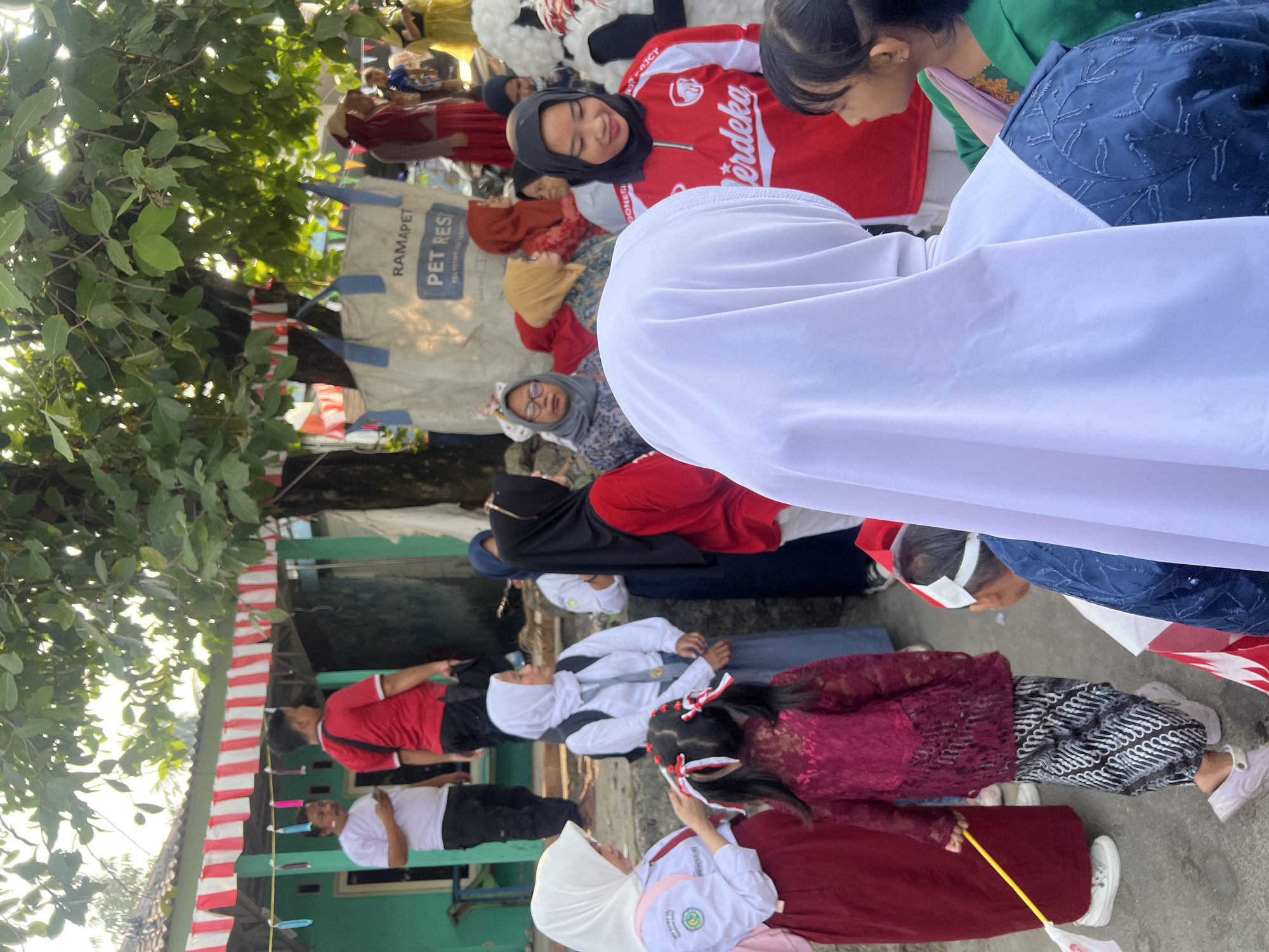 The image depicts a gathering of people, likely for a community event or celebration, in Klapanunggal, Indonesia. In the foreground, a person wearing a white hijab is facing away from the camera, with their back to the viewer. To their right, a young girl with dark hair and wearing a green jacket is looking to the left. Behind the person in the white hijab, a woman in a red shirt with "Merdeka" written on it is smiling. Other individuals are visible in the background, including people in traditional clothing and school uniforms, some of whom are wearing hijabs.
The environment suggests an outdoor setting in a residential area. A building with a green facade and a white-tiled roof is visible on the left. Red and white striped banners are hung across the front of the building and strung between trees, indicating a festive occasion, possibly related to Indonesian Independence Day. A large white bag with blue lettering that reads "RAMAPET PET RES" is also present, along with trees providing shade. The ground is a paved or dirt surface. The overall impression is one of a lively community event taking place during daylight hours.