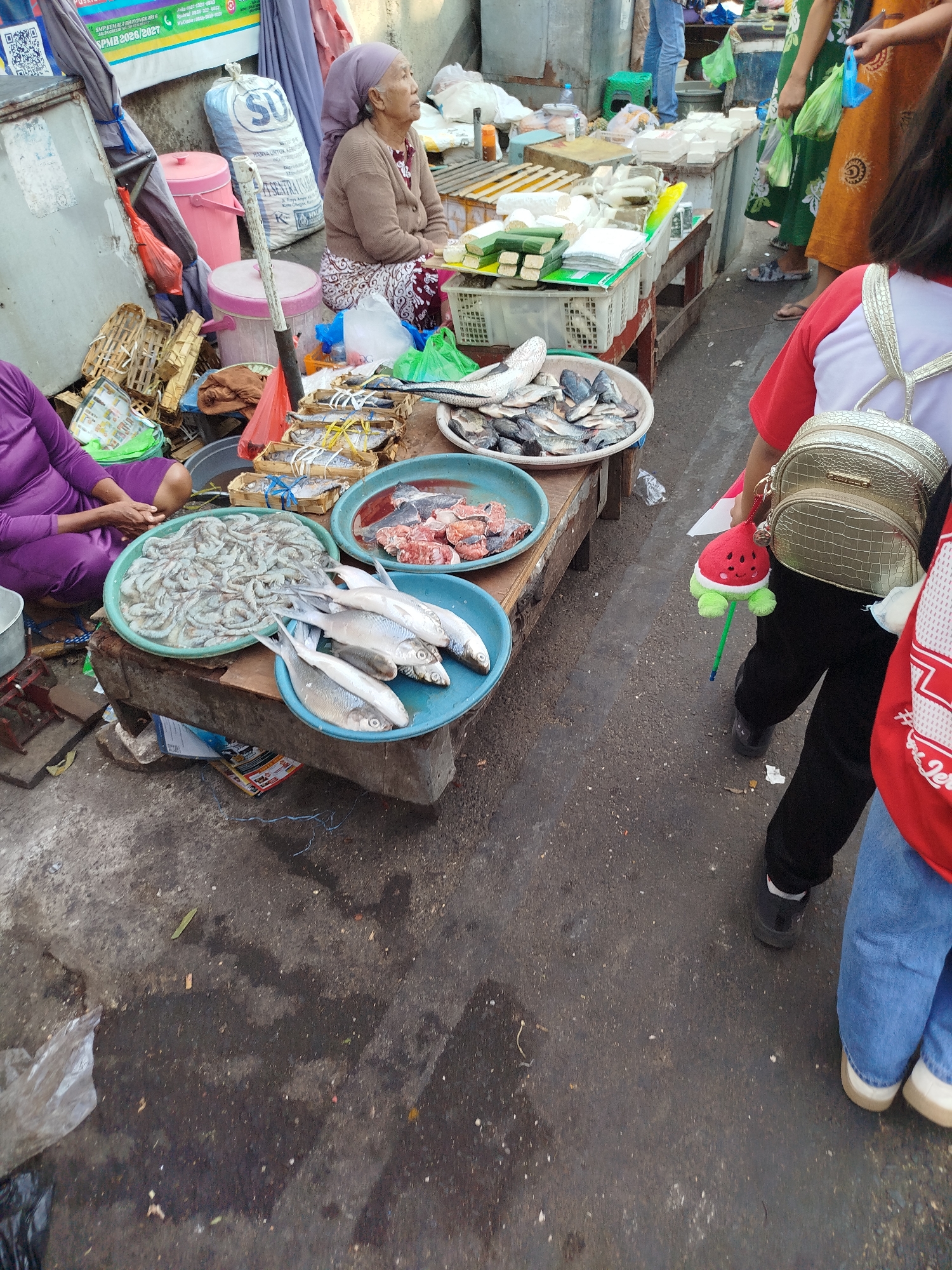 An outdoor market scene in Surabaya, Indonesia, displays various food items for sale. In the foreground, a person wearing a purple garment sits next to a large green tub filled with shrimp. Adjacent to this, a blue bowl holds several silver-colored fish. Another blue bowl to the right contains pieces of raw meat. Behind these, numerous small baskets, some containing fish, are arranged on a wooden table. Further back, an elderly woman wearing a headscarf and brown cardigan sits on the ground. To her left are pink buckets and a large white bag. Beyond the stalls, people can be seen standing and shopping. The ground is paved with asphalt, and scattered debris is visible.