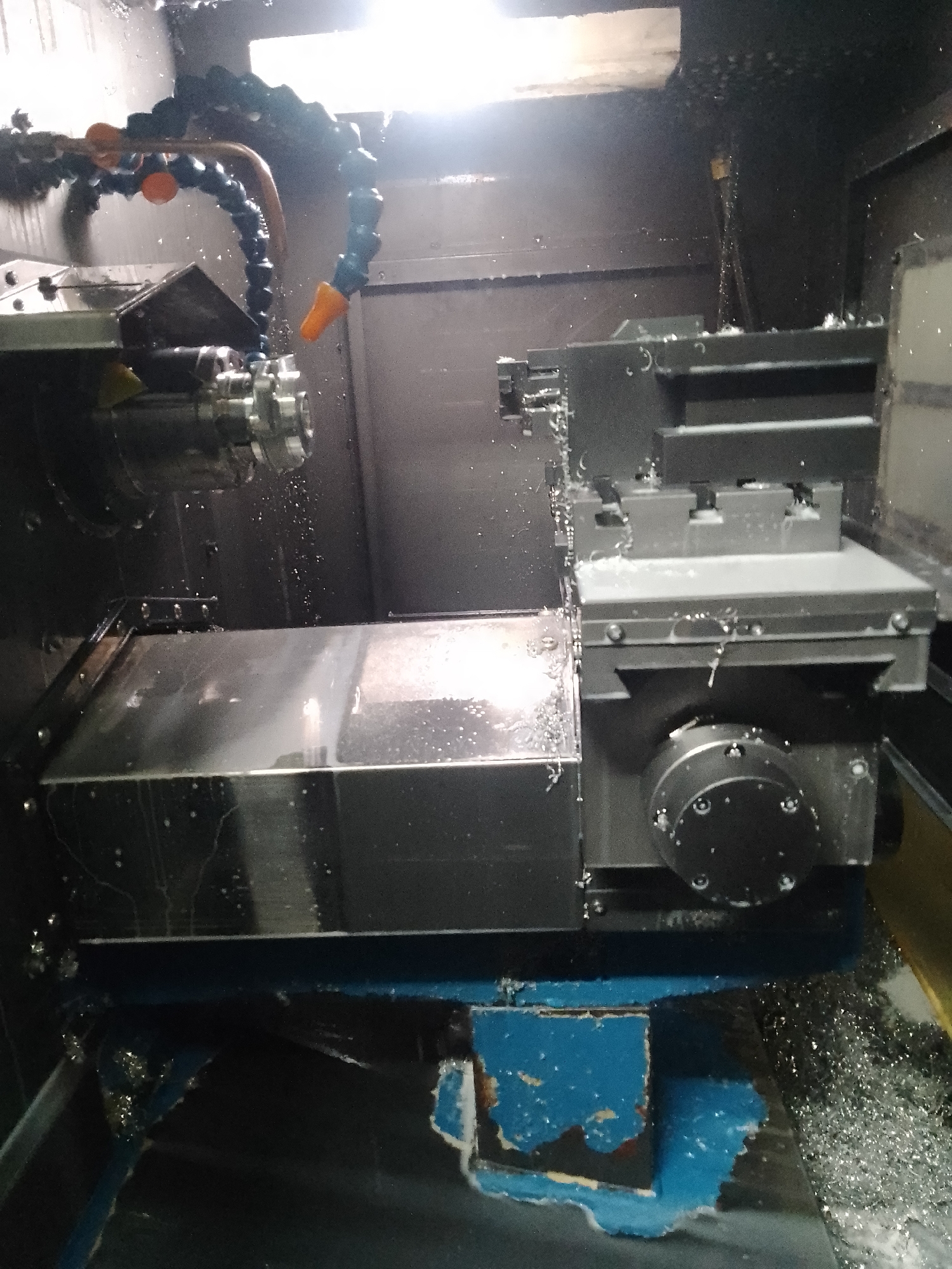 This media file shows a close-up view of a metal-cutting machine, likely a CNC lathe, in operation. The scene is inside a workshop, suggested by the industrial machinery and the presence of metal shavings. A coolant hose is visible, spraying a liquid onto the workpiece and cutting tool to keep them cool during the machining process. The machine itself appears to be made of dark metal and features various components such as a chuck, a cutting tool holder, and a movable carriage. Metal shavings are scattered around the base of the machine, indicating that material is actively being removed from a workpiece. The lighting suggests an indoor, possibly artificial, light source, and the overall impression is one of active manufacturing. There is no visible text in the image. The location context provided is Ciantra, Indonesia, which places this industrial activity in a specific geographical setting.