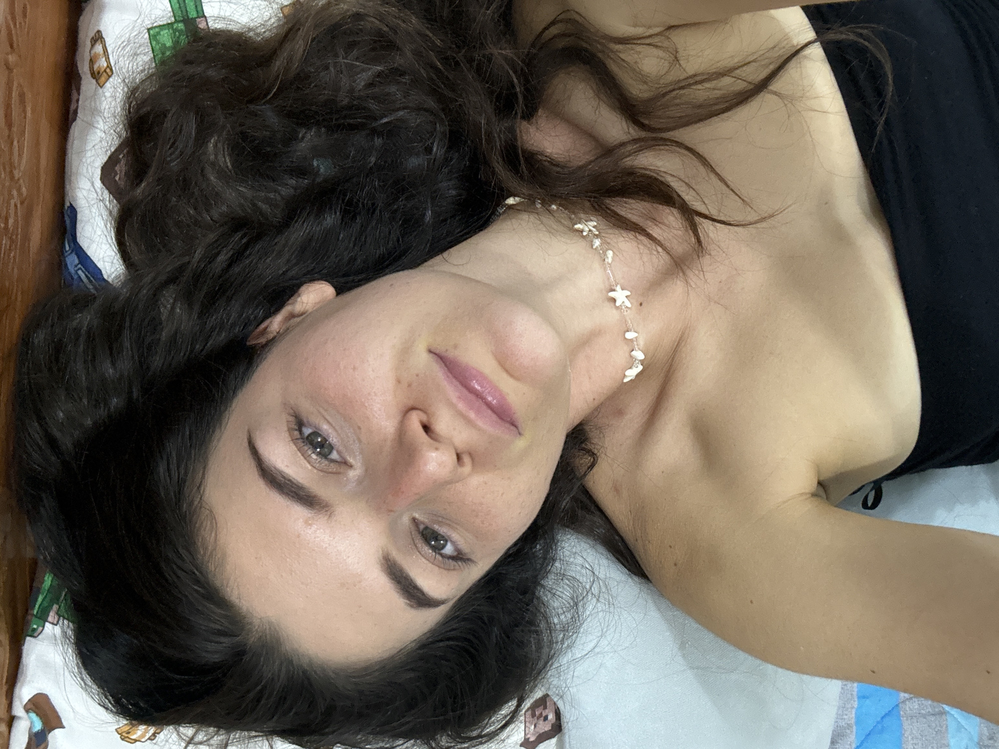 A woman with dark, wavy hair is lying down, likely on a bed, in what appears to be an indoor setting. She has fair skin, freckles on her face, and light-colored eyes. She is wearing a black top and a necklace with starfish-shaped elements. The pillow under her head has a patterned design. The location is likely a bedroom in a private residence, given the personal items present. There is no information to suggest this scene is located specifically in Mũi Né, Vietnam or any other specific city.