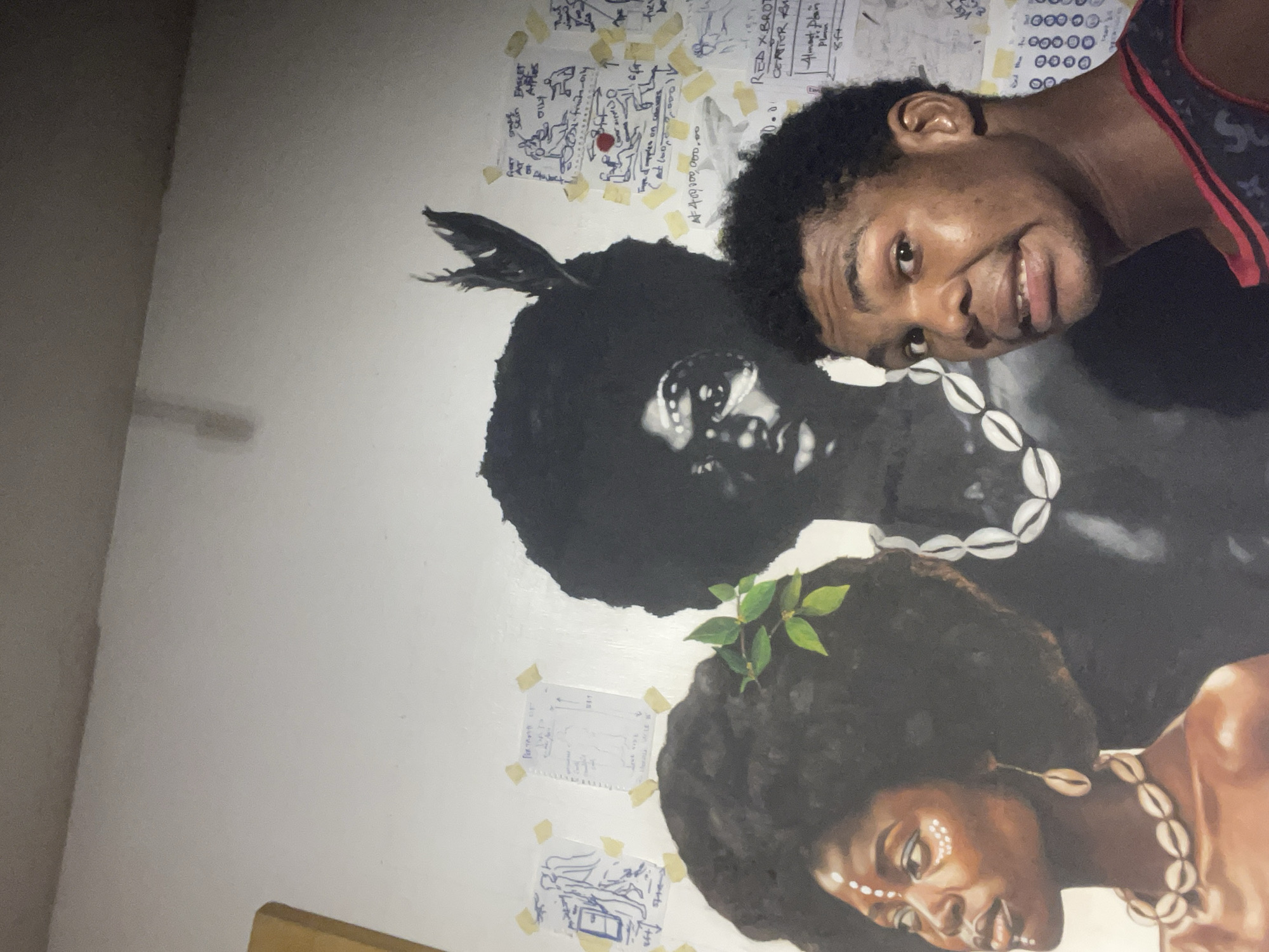 In Ado, Nigeria, a young Black man with dark, curly hair and light brown skin stands in front of a mural on a white wall. He is wearing a black and red patterned tank top and looking at the camera with a slightly bewildered expression. The mural features three figures. On the left, a woman with dark skin, a large afro, and shell jewelry is depicted in profile. She has white face paint markings. In the center, a man's face with a prominent afro and a feather headdress is shown. To the right of the central figure, another afro is visible. The wall behind the mural is covered with various sketches and notes, some of which appear to be in English. The lighting suggests an indoor setting, possibly a studio or gallery.