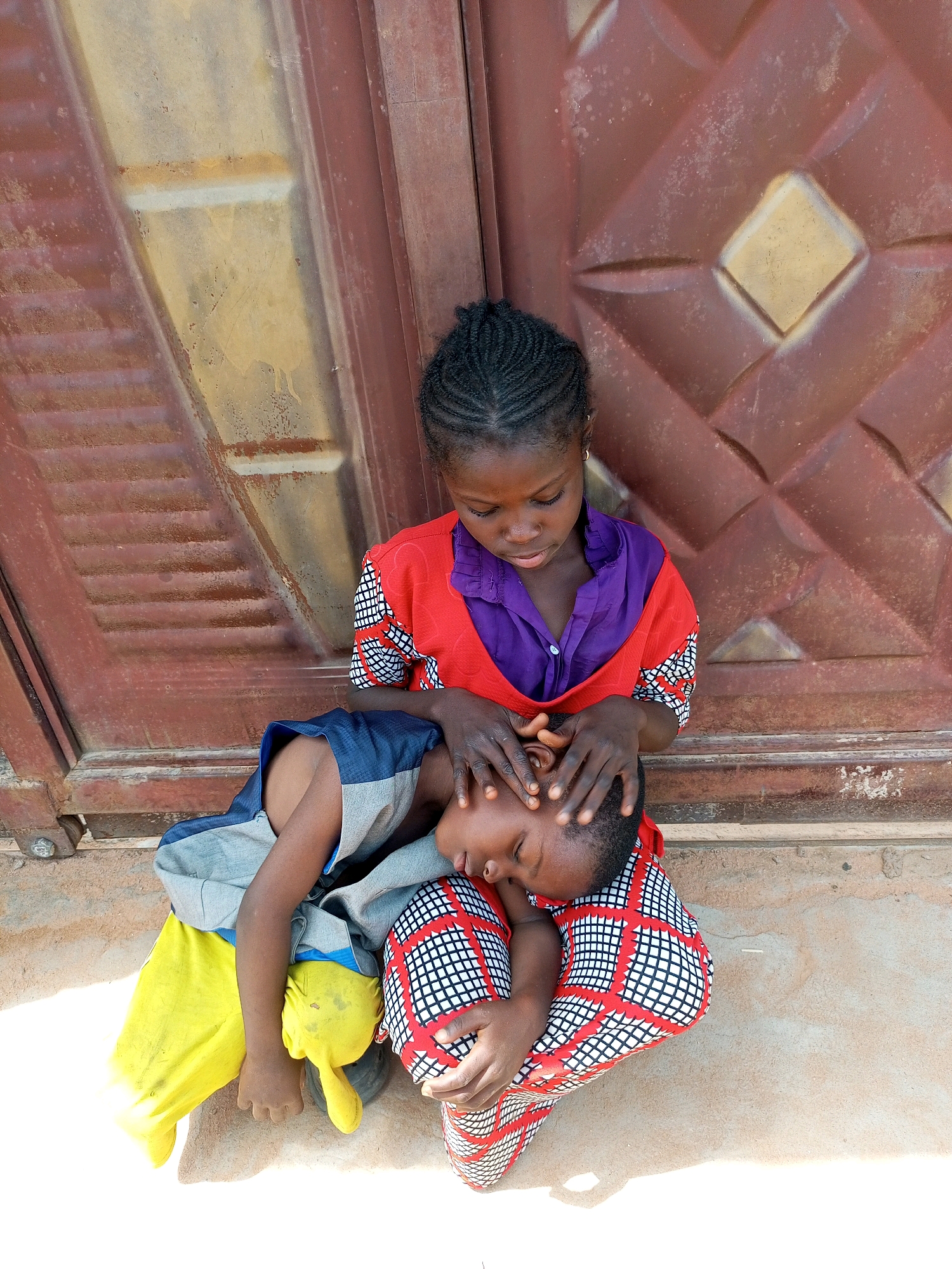 A young girl with braided hair sits with her eyes downcast, her hands gently resting on the head of a younger child who is asleep on her lap. The younger child is wearing a blue and gray shirt with yellow pants. The girl is wearing a red and purple shirt with a patterned skirt. They are sitting in front of a weathered, red metal door with a diamond pattern. The ground is sandy and appears to be outdoors. The lighting suggests it is daytime. This scene is located in Tempure, Nigeria.