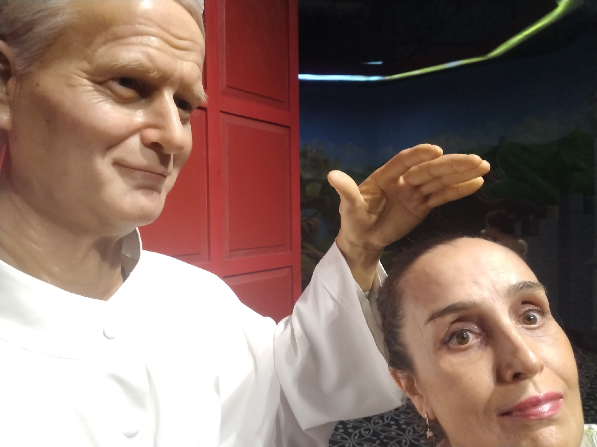 The image shows a woman posing with a wax figure of Pope John Paul II. The Pope is wearing a white shirt and has a slightly smiling expression. The woman's hand is raised and slightly touching the head of the Pope, as if she's receiving a blessing. The background includes a red wall and a painted scene of a landscape. The location is likely a wax museum.