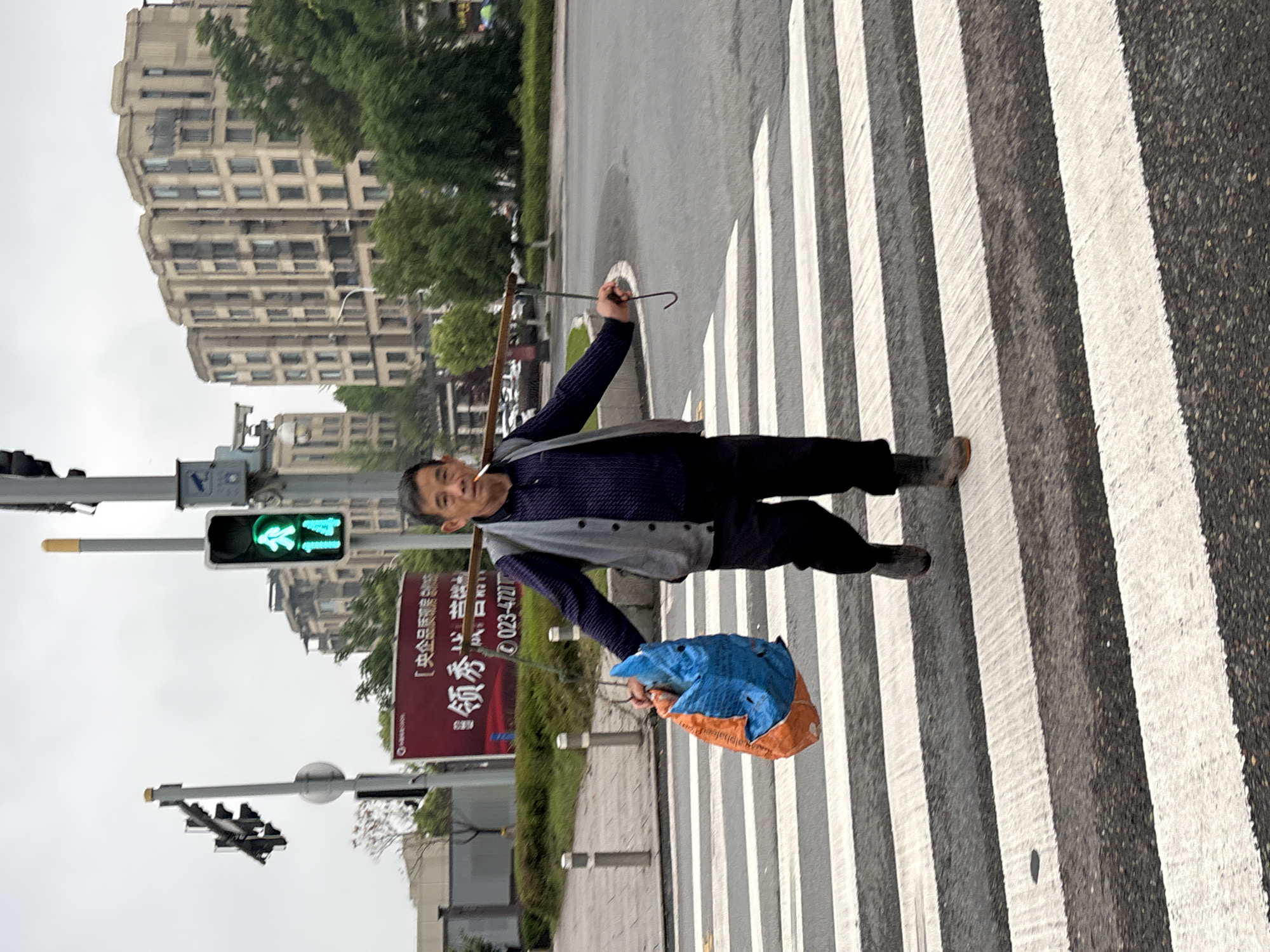A man is walking across a pedestrian crossing in Jiangjin, China. He is carrying a blue and orange bag in his left hand and a walking stick over his shoulder. The pedestrian light shows green. In the background, there is a sign written in Chinese and a large building. The road surface is wet.