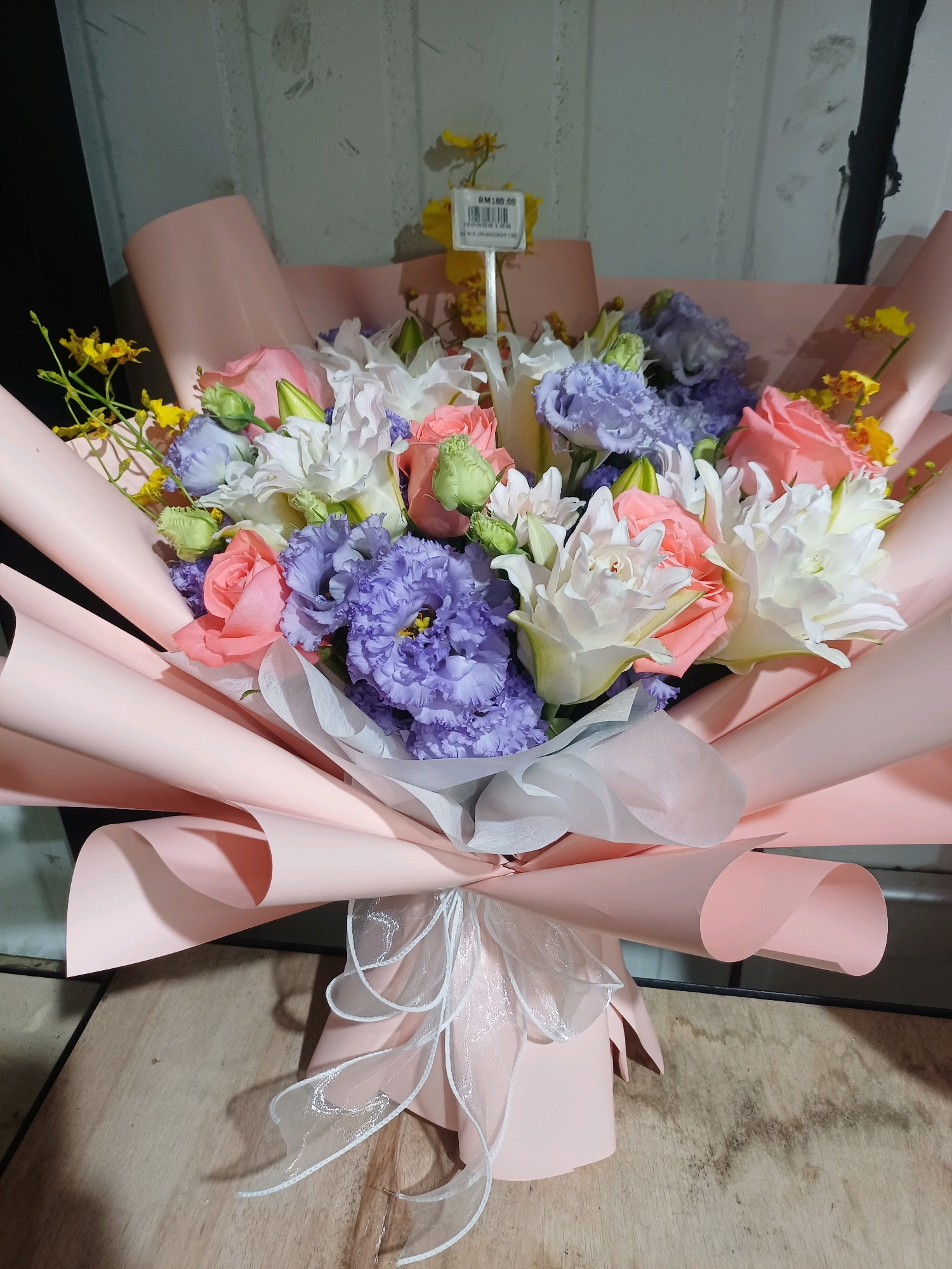 The image shows a bouquet of flowers wrapped in pink paper, tied with a white ribbon. The bouquet is placed on a wooden surface, possibly a table or counter. In the background, there is a grey wall with a textured pattern. A price tag with "RM 180.00" is visible among the flowers. The scene appears to be indoors, in a setting that could be a florist shop or a personal space in Kuala Lumpur, Malaysia. The lighting is artificial, creating shadows on the wooden surface. There are no people or animals visible in the image. The bouquet contains a variety of flowers, including pink roses, purple lisianthus, and white lilies, along with some small yellow flowers. The overall presentation is neat and colorful.