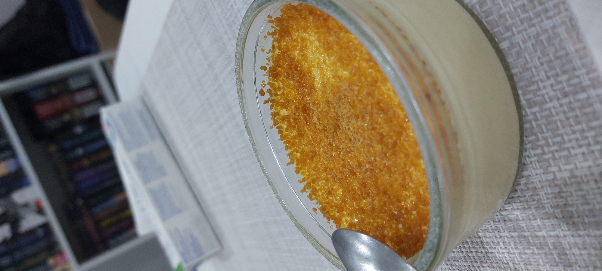 A close-up, tilted overhead shot depicts a small glass dish containing a dessert, potentially crème brûlée, resting on a woven, light-colored placemat. The dessert has a light yellow custard base topped with a golden-brown caramelized sugar crust. A spoon is partially visible at the bottom left corner of the dish. The background is blurred but shows a white table surface, part of a white bookshelf filled with books, and a box with blue and white text. The location is unlikely to be determined as Yagodovo, Bulgaria.