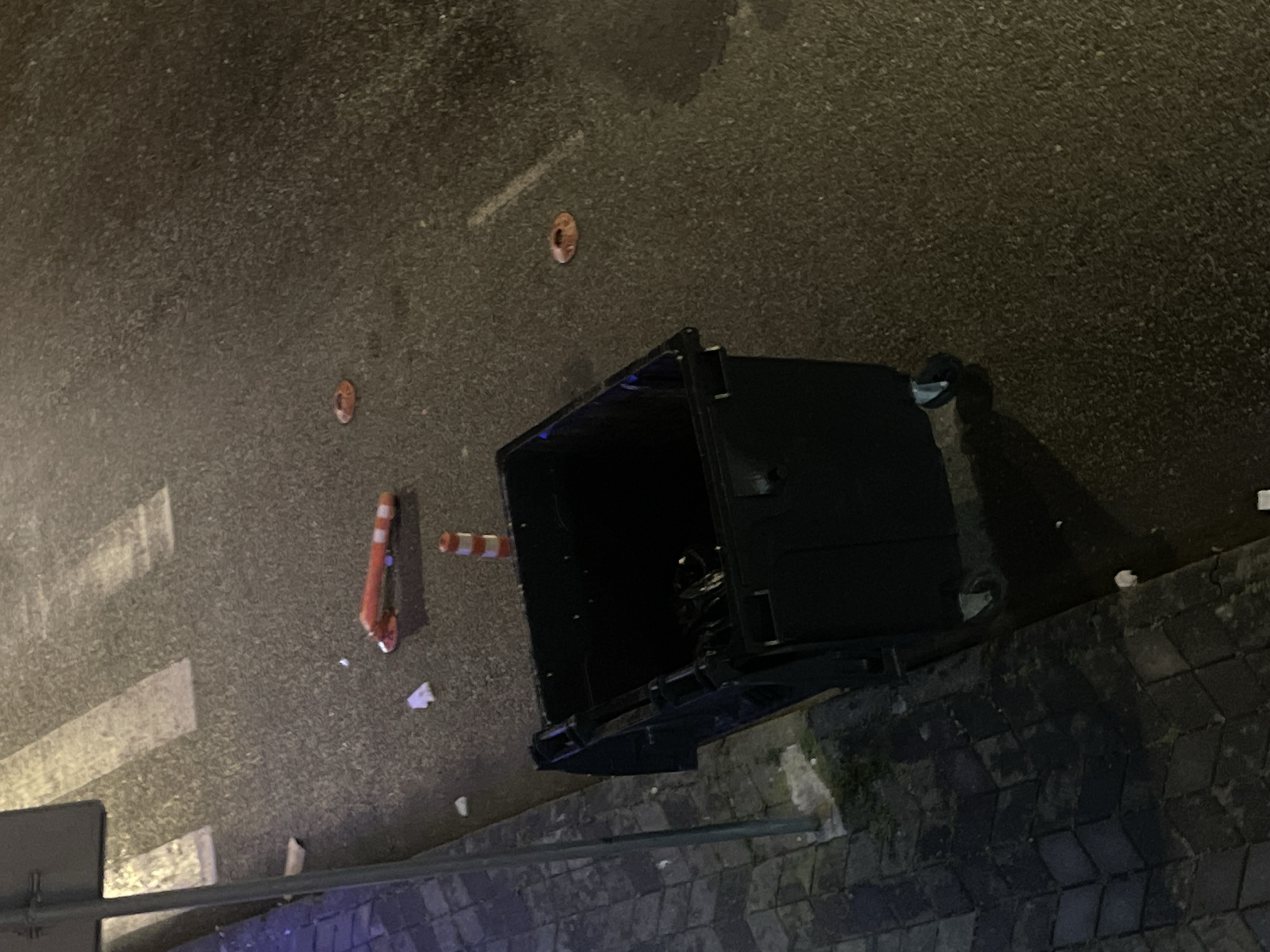 The image depicts an outdoor scene at night, likely on a street in Batumi, Georgia, given the context. The foreground features a large, black dumpster with wheels. The ground is asphalt, wet and reflective, suggesting recent rain or street cleaning. Several objects are scattered on the asphalt. One is a traffic cone, orange and white striped, lying horizontally. Another, partially visible, is a shorter orange and white striped traffic cone standing upright. There are also two round, flat, orange objects on the ground, possibly discarded food wrappers or lids. White paper debris is visible near the horizontal traffic cone and near the edge of the paved sidewalk on the left. The sidewalk itself is made of dark cobblestones arranged in a diamond pattern. A lamppost or pole is visible on the far left, with a dark rectangular sign attached to it. The overall impression is of a slightly untidy urban street scene at night.