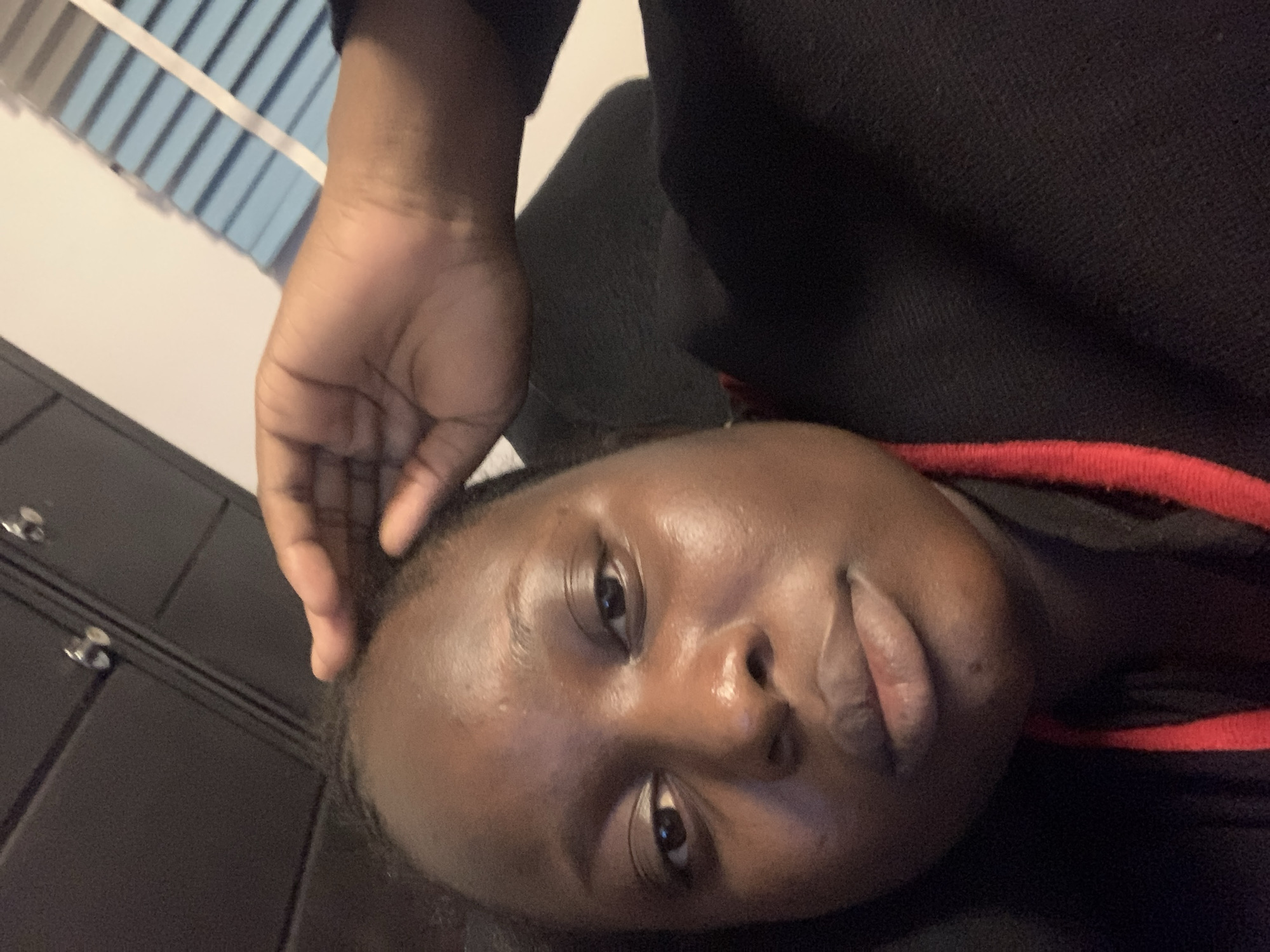 A close-up selfie of a Black woman is shown, with her hand resting on her forehead. The woman has dark skin, brown eyes, and full lips. She is wearing a black top with red trim. The background of the image appears to be an indoor setting with dark cabinetry and blinds. The lighting is dim, casting shadows on the woman's face. The image is not associated with a specific location in the city of Unknown, Unknown.