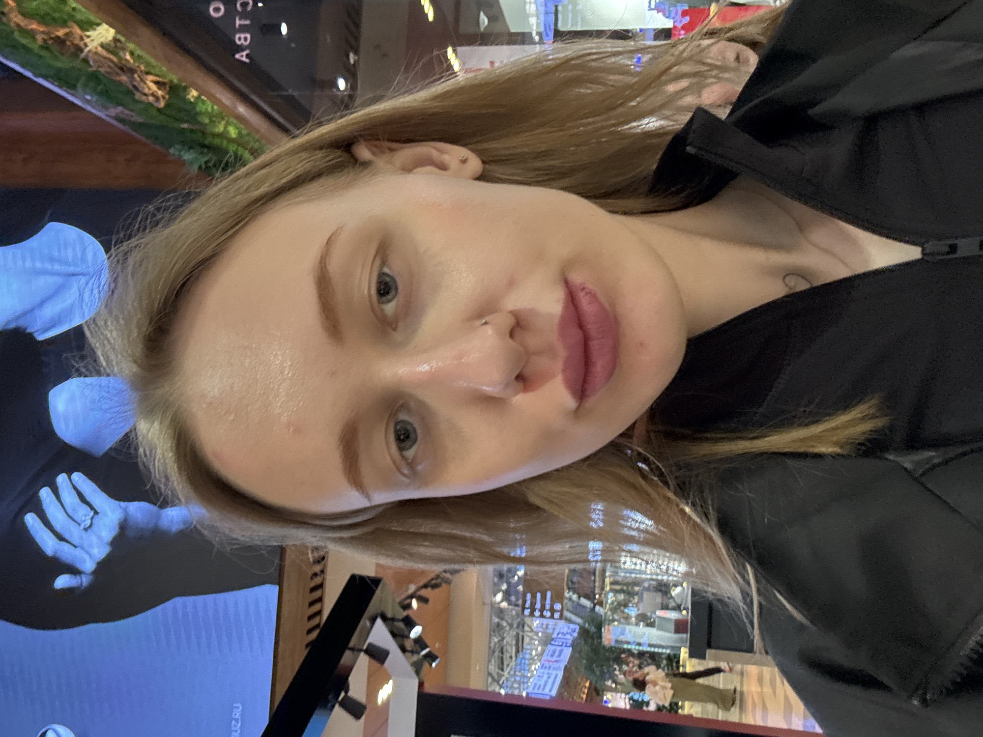 A selfie of a fair-skinned woman with blonde hair, blue eyes, and a nose piercing. She is wearing a black jacket. A tattoo is visible on her chest. The background appears to be a public space with a display screen showing an image of a hand. Other displays and interior architecture are visible. The location is not identifiable as Ol'ginskaya, Russia based on the provided information.