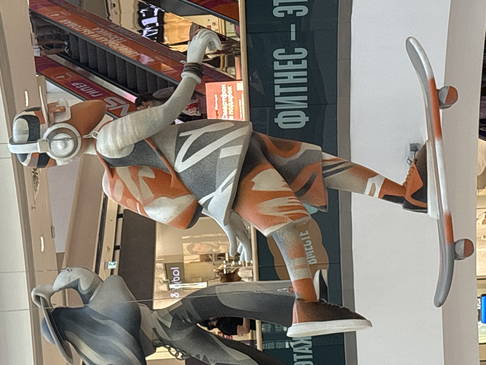 The image depicts a large statue of a person on a skateboard inside a shopping mall. The statue is colorful, with orange and gray as prominent colors. The person is wearing headphones and a backpack. In the background, there are escalators, signs in Russian, and other shoppers. One sign reads "ФИТНЕС" which translates to "FITNESS." The overall environment suggests a modern commercial space. It is a photograph taken in the city of Novosibirsk, Russia.