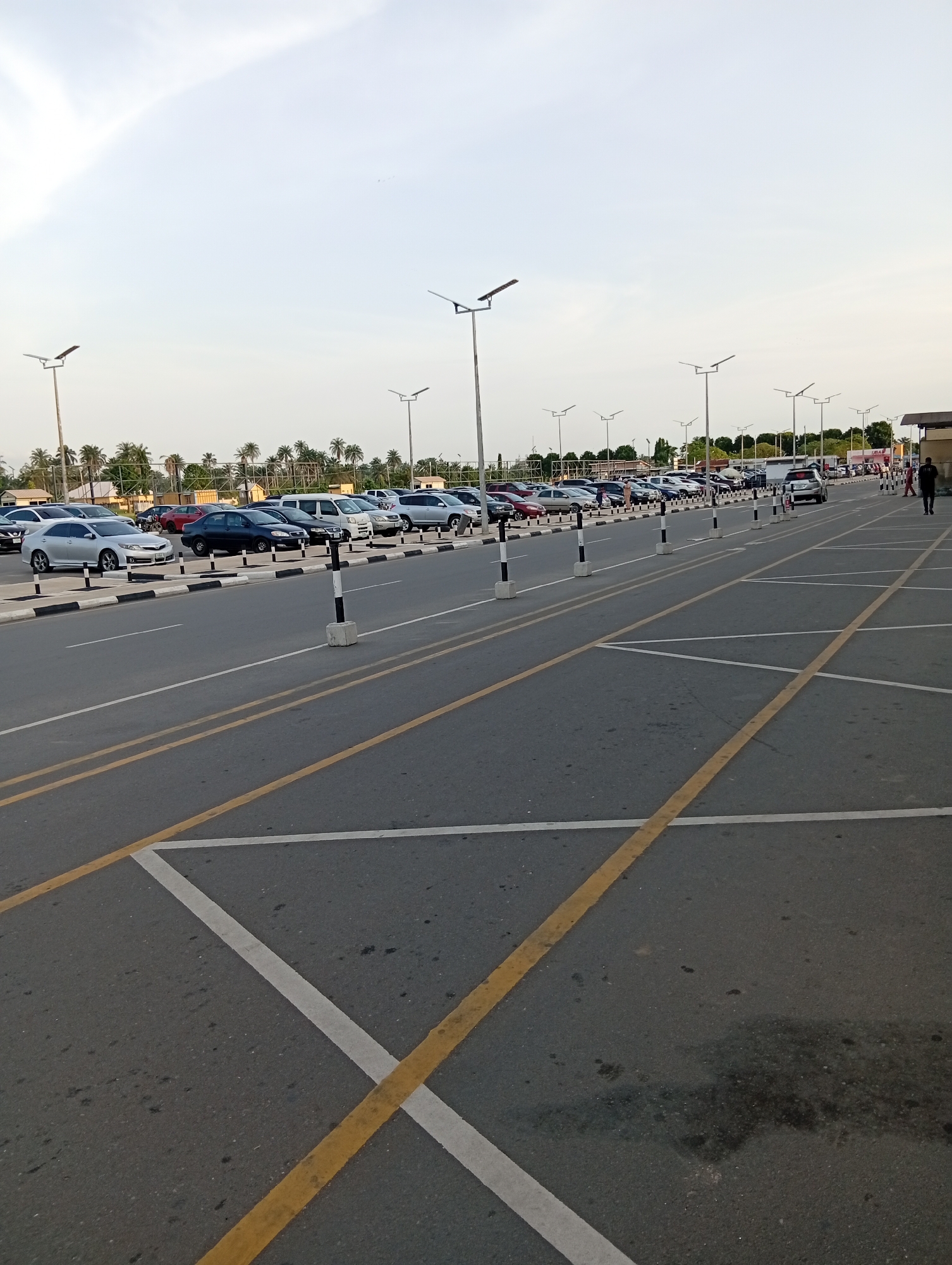 The image shows a large, mostly empty parking lot in Omagwa, Nigeria. Rows of parked cars are visible in the background, separated from the empty parking spaces by a row of bollards. Overhead are tall lampposts. The parking spaces are demarcated with yellow and white lines on the asphalt. A person is walking in the background near a building. The sky is overcast.