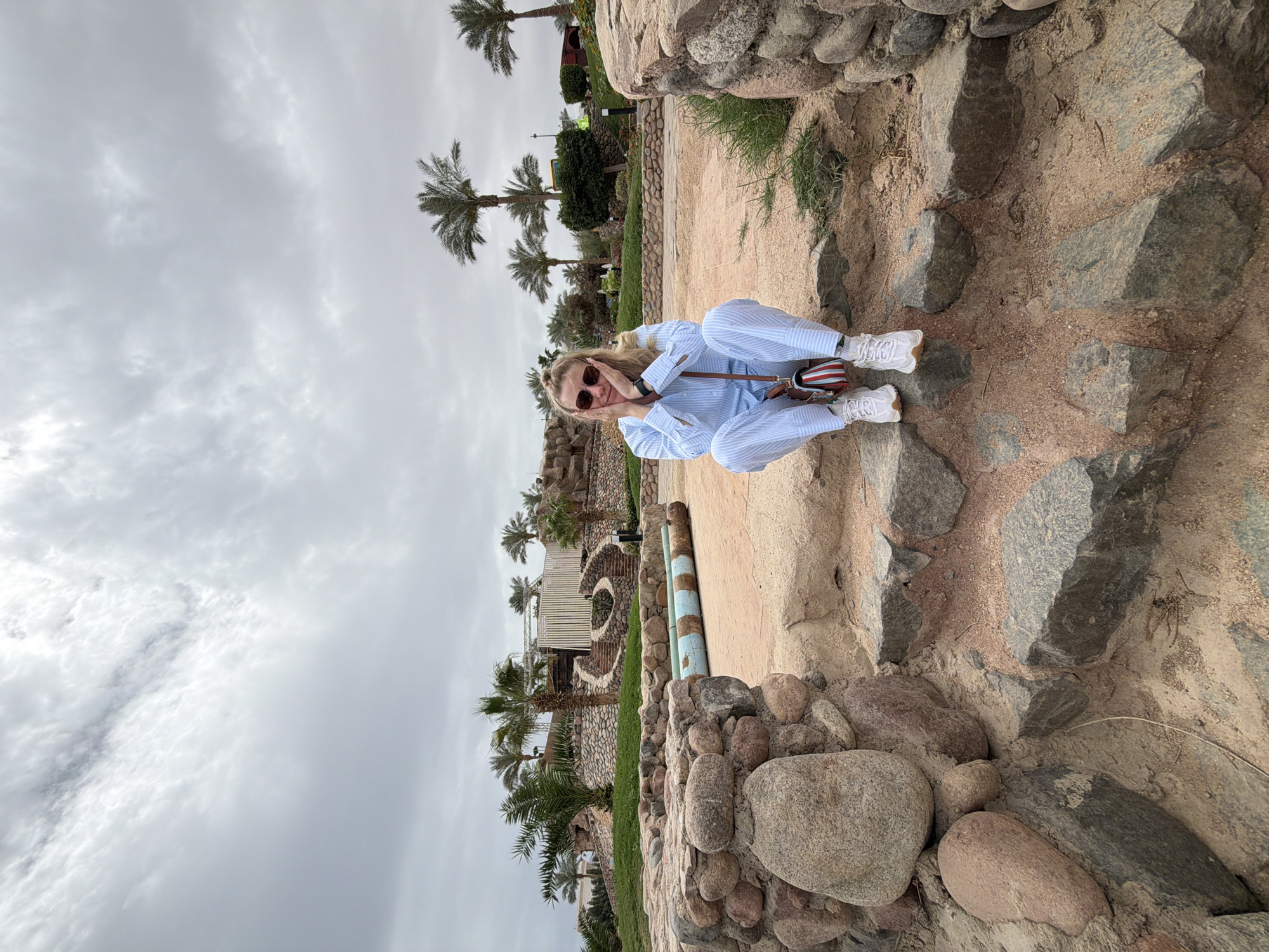 A woman with blond hair wearing a blue and white striped outfit and red sunglasses is squatting on rocks in Sharm El Sheikh, Egypt. She is wearing white sneakers and has a striped bag over her shoulder. Palm trees and buildings are visible in the background under a cloudy sky. The ground is sandy and rocky.