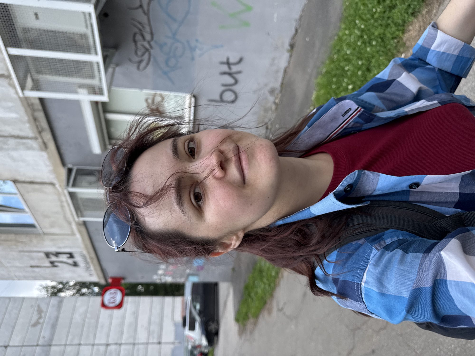 An outdoor, eye-level, medium shot captures a young woman with brown hair and brown eyes, wearing sunglasses on her head. She is looking at the camera with a slight smile. She is dressed in a red t-shirt and an open blue plaid button-up shirt, with a black backpack visible. The background shows a grey building with graffiti on the lower portion of the wall, featuring the word "but" and other markings. To the left of the building, a red circular sign with white text is visible on a pole. A sidewalk and a grassy area with scattered white flowers are in the foreground. A dark car is parked in the background on the left. The scene is likely on a street in Izhevsk, Russia, indicated by the building's architecture and the graffiti style.
