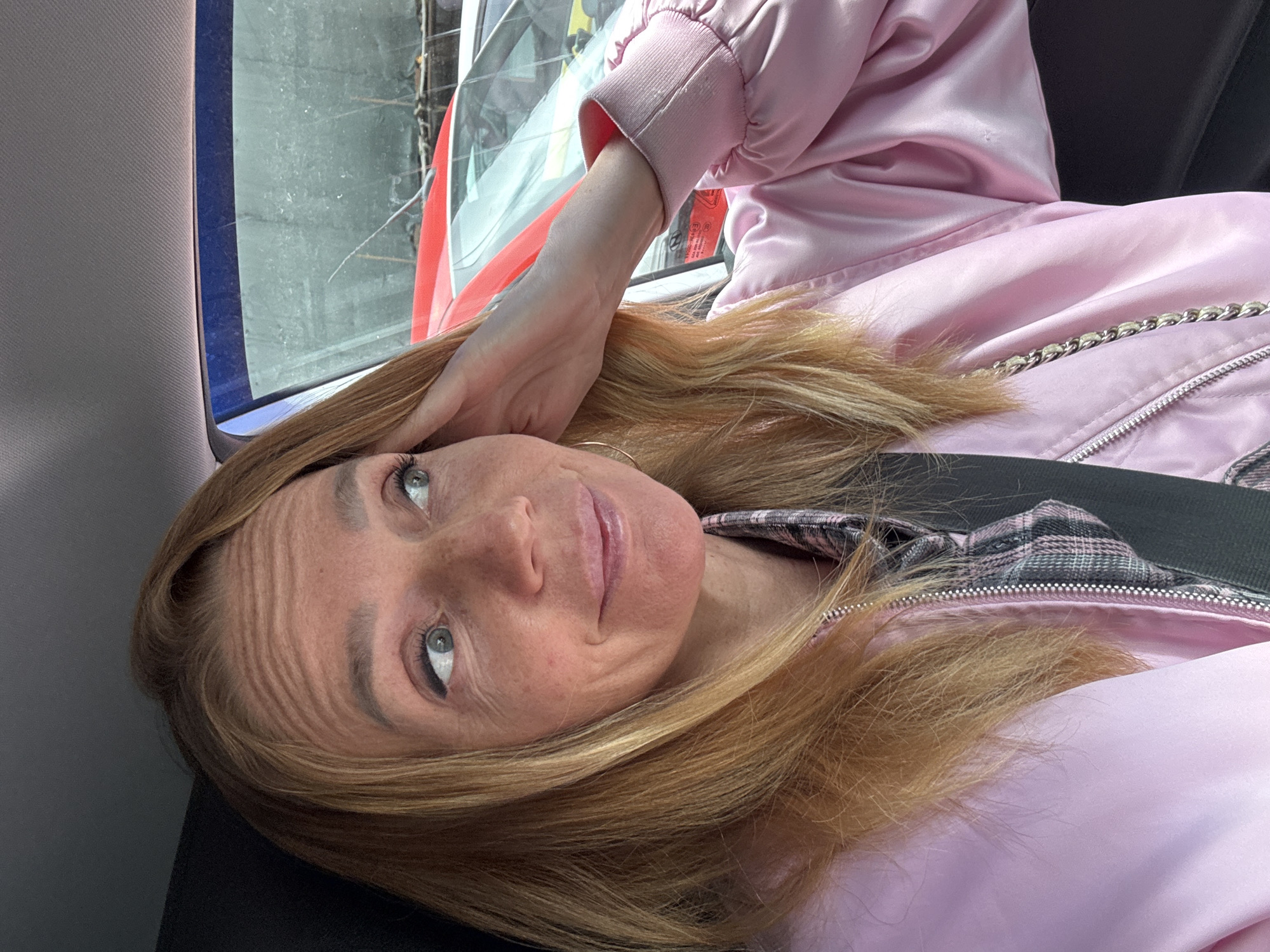 A woman with blonde hair, wearing a pink bomber jacket over a plaid shirt, is seated in the driver's seat of a car. Her left hand is raised to her forehead, and she is looking to her right. The interior of the car is visible, as is the windshield and the exteriors of two cars parked behind. The scene appears to be in a parking lot.