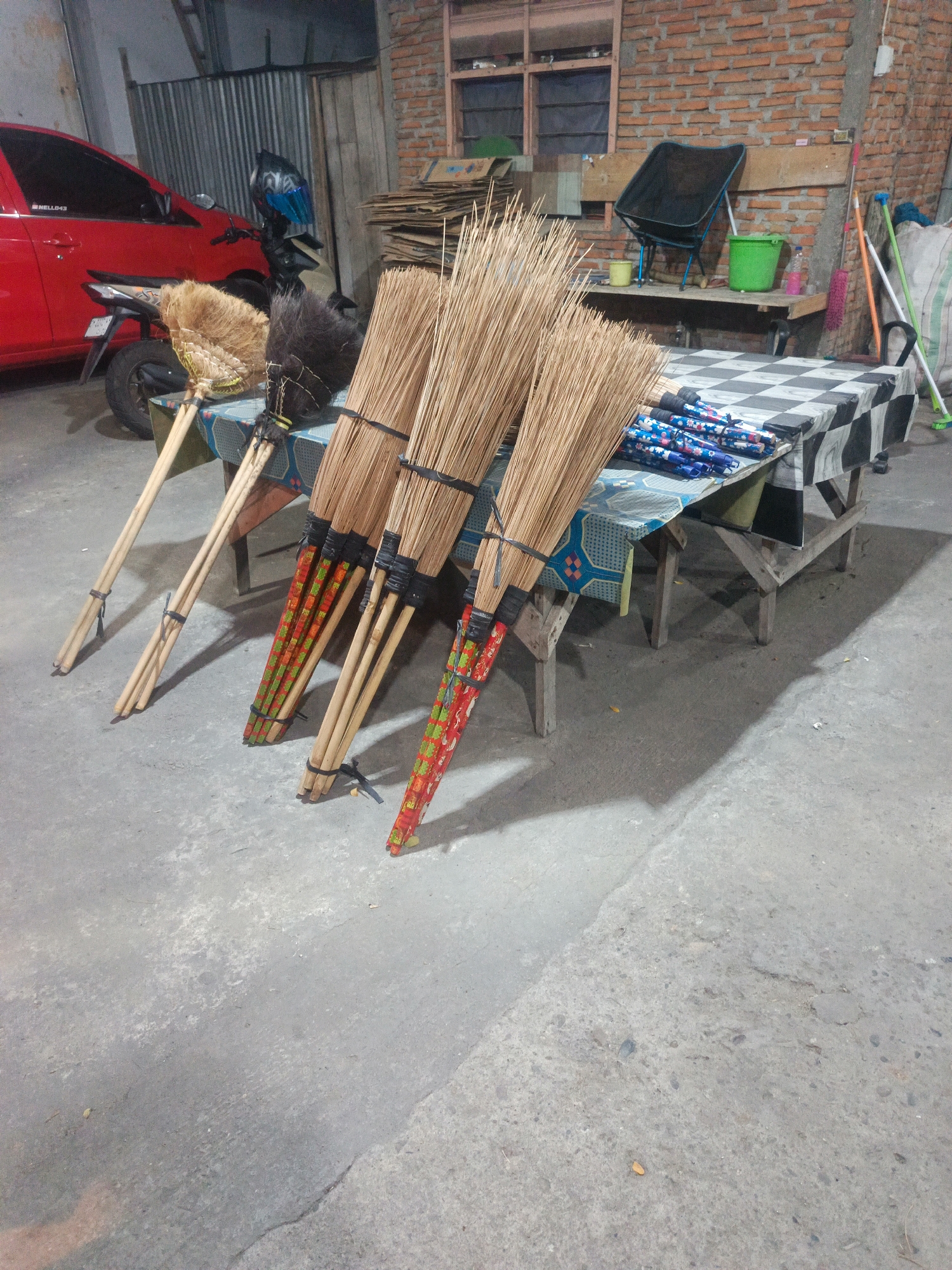 The image depicts a dimly lit outdoor scene, likely a marketplace or a vendor's stall, in Ulapato A, Indonesia. In the foreground, four brooms with natural fiber bristles and wooden handles are displayed, some adorned with colorful, patterned wrapping near the bristles. Behind the brooms, a rustic wooden table is set up, covered with two checkered cloths. On the table, several rolled-up items, possibly umbrellas or flags, are neatly arranged.
To the left, a red car is partially visible, and behind it, a motorcycle is parked with a helmet on its handlebars. Further back, a brick wall forms the backdrop, with a window and a folding chair visible. A green bucket and a few other items are placed on a shelf against the wall. To the right of the table, a large white sack and several cleaning tools, including mops with colorful handles, are leaning against the wall. The ground is a concrete surface, showing signs of wear and tear, with some shadows cast from the objects in the scene. The overall impression is that of a humble, functional setup for selling goods.