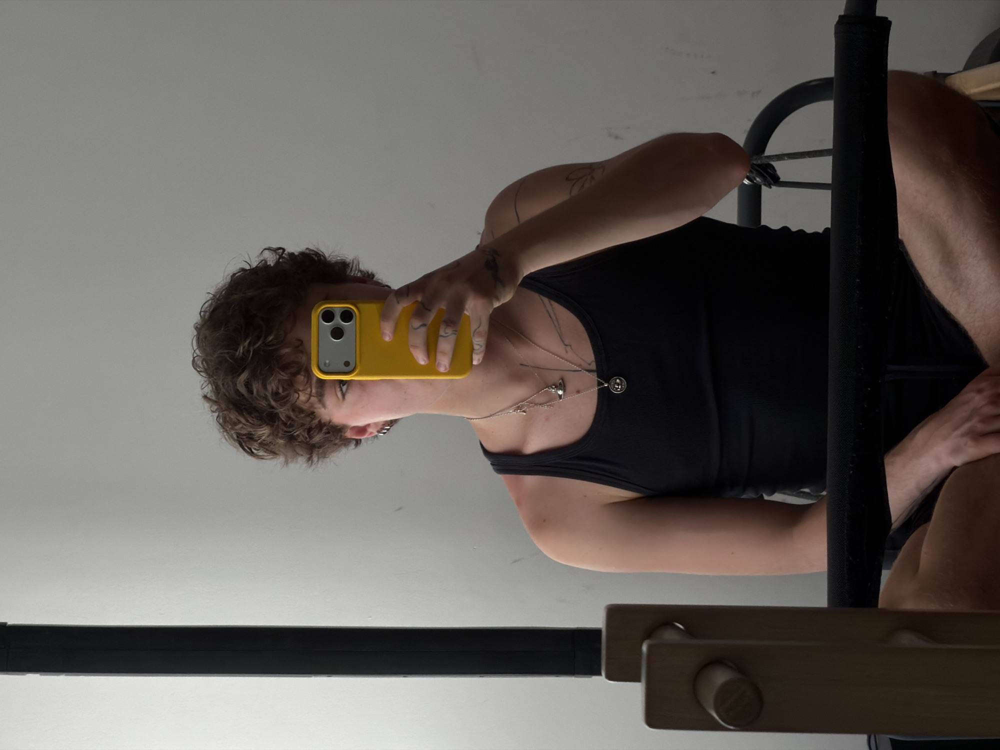 A person is taking a selfie in a mirror. The person has curly brown hair, is wearing a black tank top, and holding a yellow phone. The person appears to have tattoos on their arms. They are wearing multiple necklaces. The mirror is reflecting the room, which includes a black metal chair and a wooden structure. The location is impossible to determine from the image.