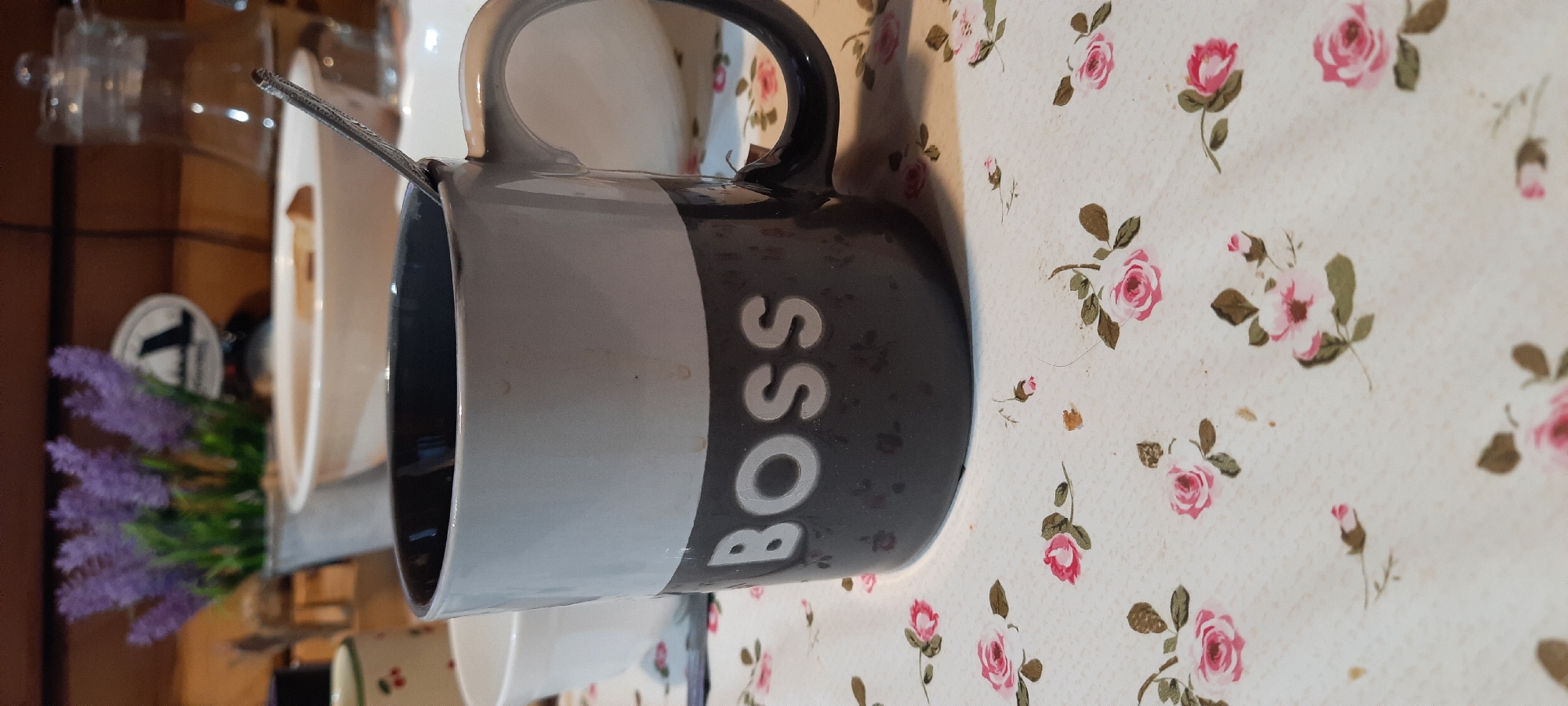 A close-up shot focuses on a dark gray mug with the word "BOSS" emblazoned in white letters. The mug is two-toned, with the upper portion being a lighter gray. A silver spoon is partially visible inside the mug, resting against the rim. The mug sits on a white tablecloth adorned with a pattern of small pink roses and green leaves. In the blurred background, a glass pitcher filled with liquid can be seen, along with a white bowl, and a vase with purple flowers. The setting appears to be indoors, possibly a home or cafe, with wooden elements visible in the background.
