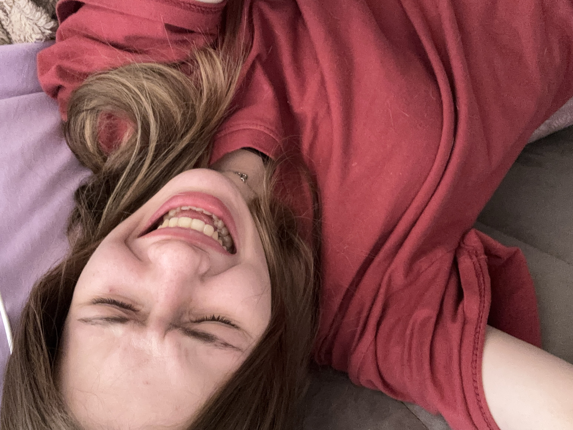 A woman lies on her back, her head tilted to the left. Her eyes are closed, and her mouth is open in a wide grin, revealing braces on her teeth. Her hair, a mix of brown and blonde, is spread around her head. She is wearing a red t-shirt. The background is a blur of purple and grey fabric. The scene appears to be indoors, possibly a bedroom or living room.