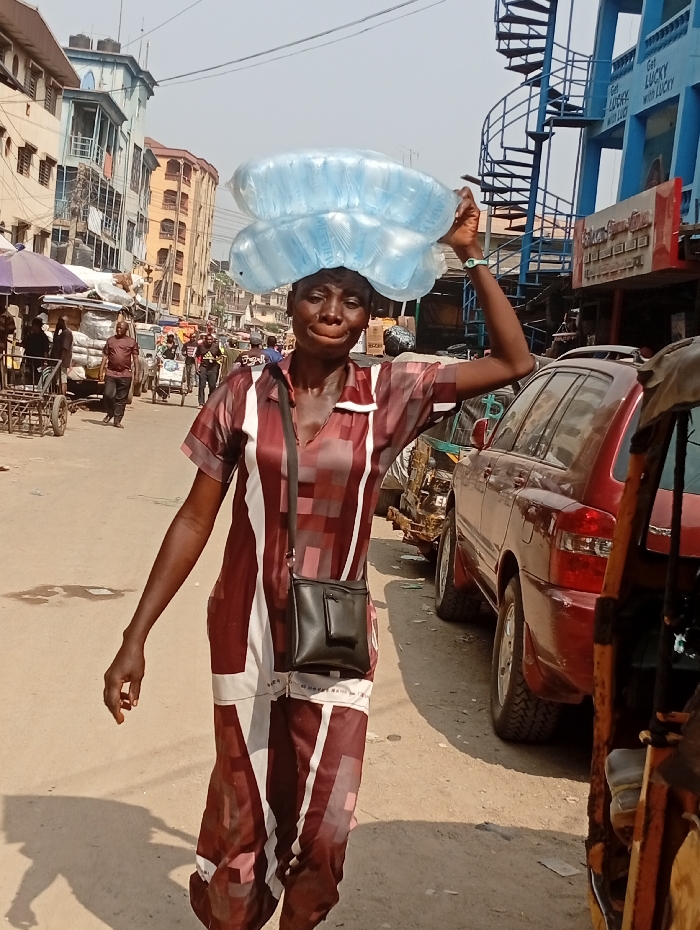 A woman carries two stacks of water sachets on her head in a bustling outdoor environment. She wears a long, patterned burgundy and white dress and a black crossbody bag. Buildings and vehicles line the street, suggesting a busy market or commercial area. The background includes people, carts, and a blue building with a spiral staircase. The scene is well-lit, capturing the everyday life and work of the woman.