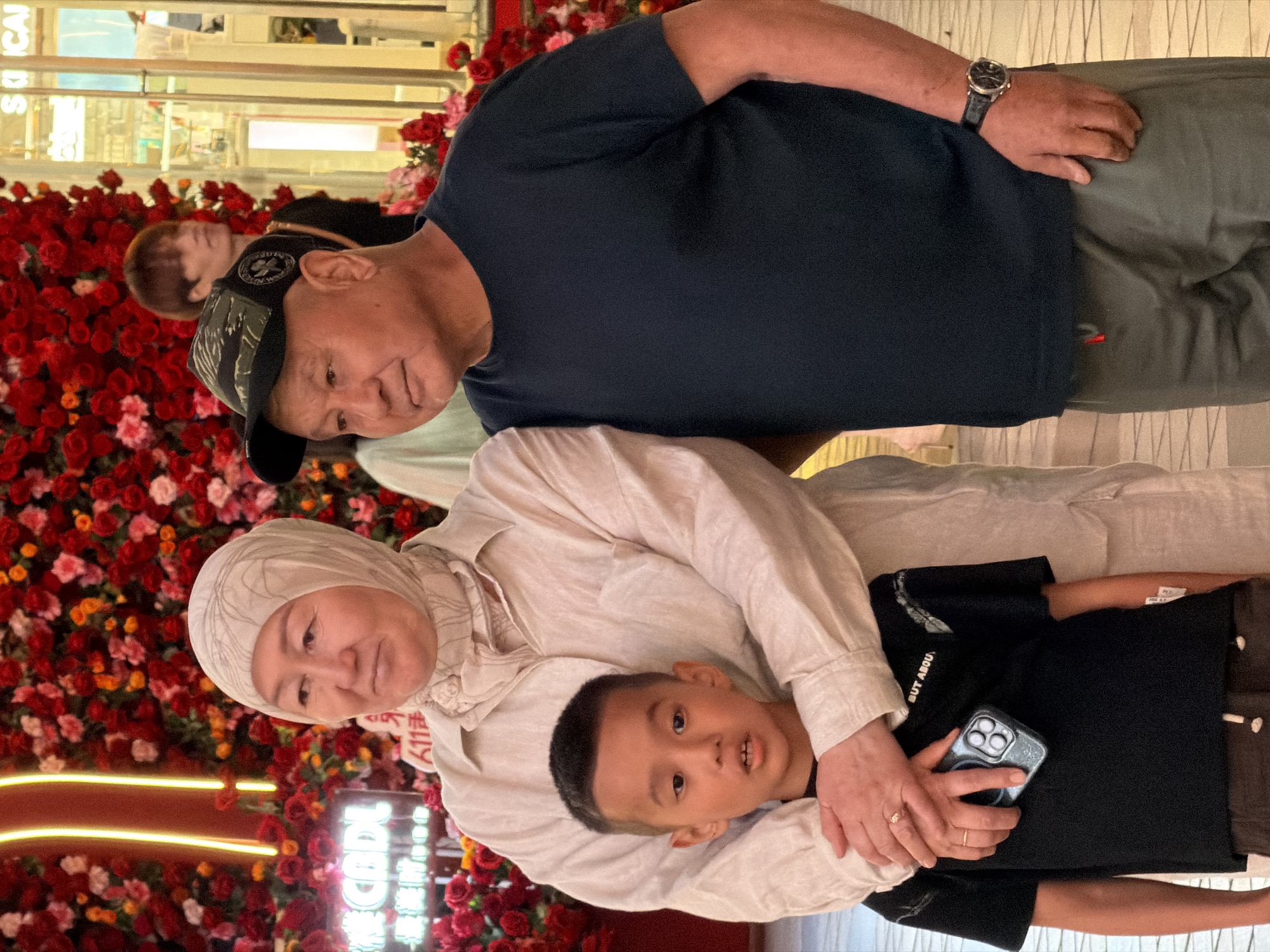 A family of three, two adults and a child, pose for a photograph in front of a large floral display of red and pink flowers, possibly in a shopping mall. The woman, on the left, wears a light-colored headscarf and a matching long-sleeved shirt. Her arms are around the boy in the center, who is wearing a black t-shirt and holding a smartphone. The man, on the right, wears a dark t-shirt, a camouflage baseball cap, and dark green pants. He has a watch on his left wrist. In the background, neon lights and a storefront sign with Chinese characters are visible. The setting appears to be indoors, likely a commercial or entertainment venue in Yuzhong District, China.