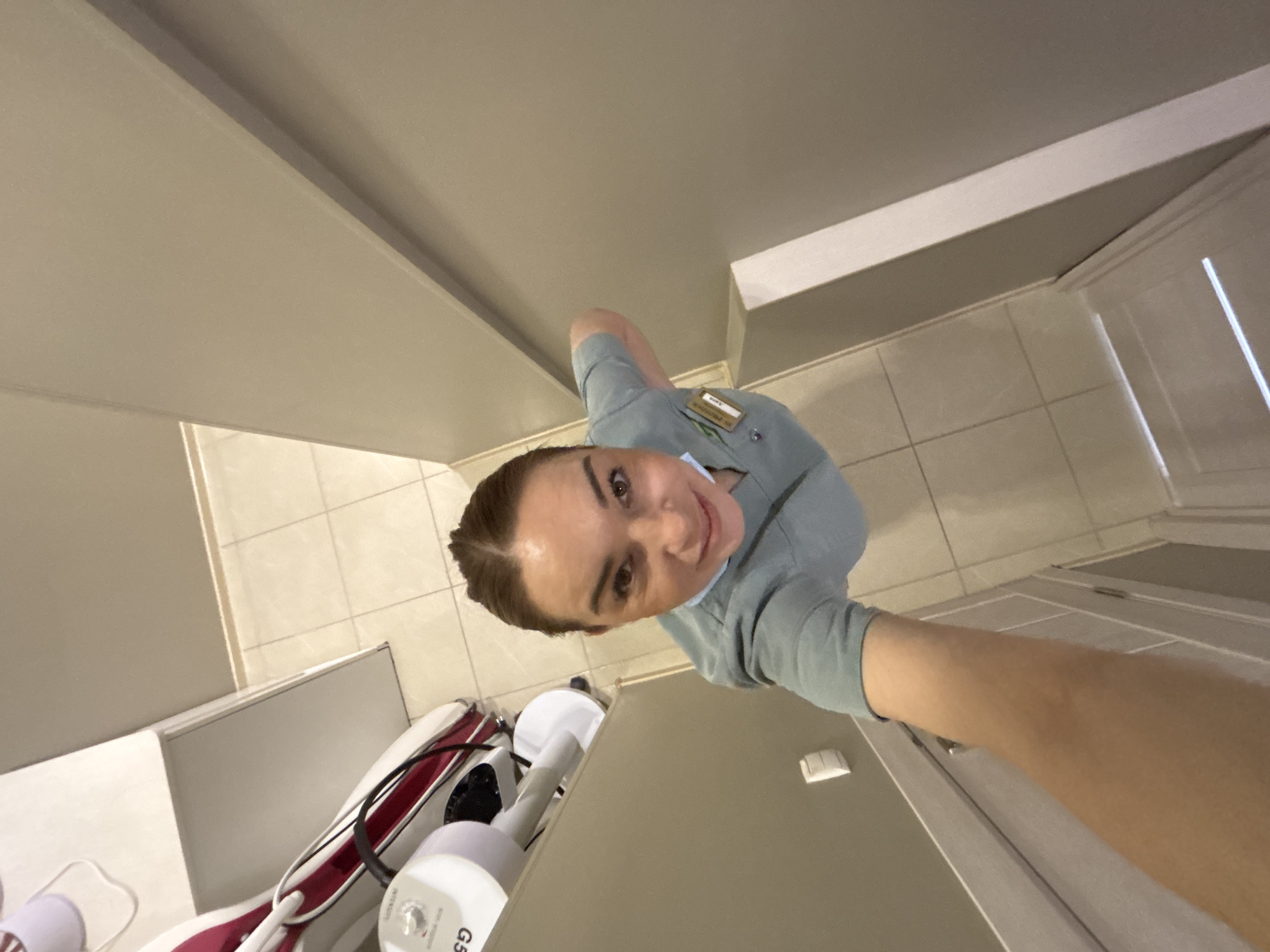 A woman in a light blue uniform takes a selfie in a room with light beige walls and tiled floors. There is a white cabinet and some medical equipment partially visible. The specific location within Vsevolozhsk, Russia, cannot be determined from the image alone.