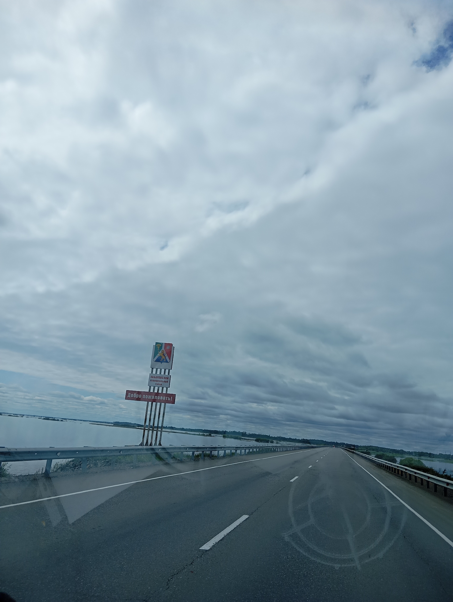 The image shows a cloudy sky over a highway with a body of water to the left. There is a sign on the side of the road that reads "Khabarovsky Municipal District" and "Welcome!". The highway appears to be a two-lane road with a guardrail on either side. The water on the left appears to be a river or lake, with trees visible on the distant shore. The sky is overcast, with thick gray clouds dominating the scene. The overall impression is one of a somewhat somber and remote landscape. The text on the sign is in Russian.