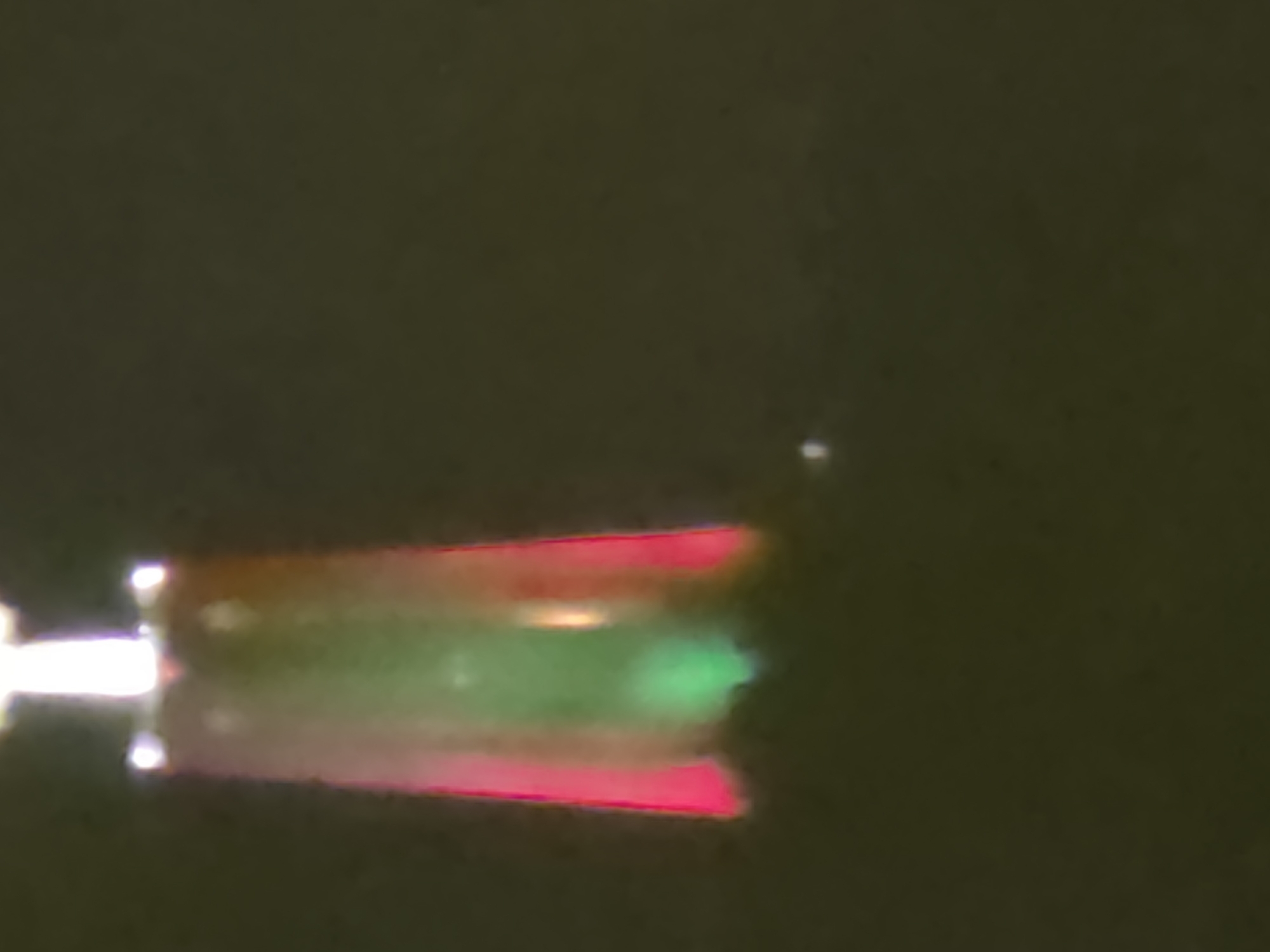 A blurred image shows a red and green illuminated object against a dark background. The object is rectangular and appears to be a light source or a display. The illumination is strongest at the top and fades towards the bottom. There is a small, bright white speck in the lower right portion of the image, which could be a distant light or a reflection. The image does not contain any recognizable landmarks or elements that would allow for identification of a specific location in Lisbon, Portugal.