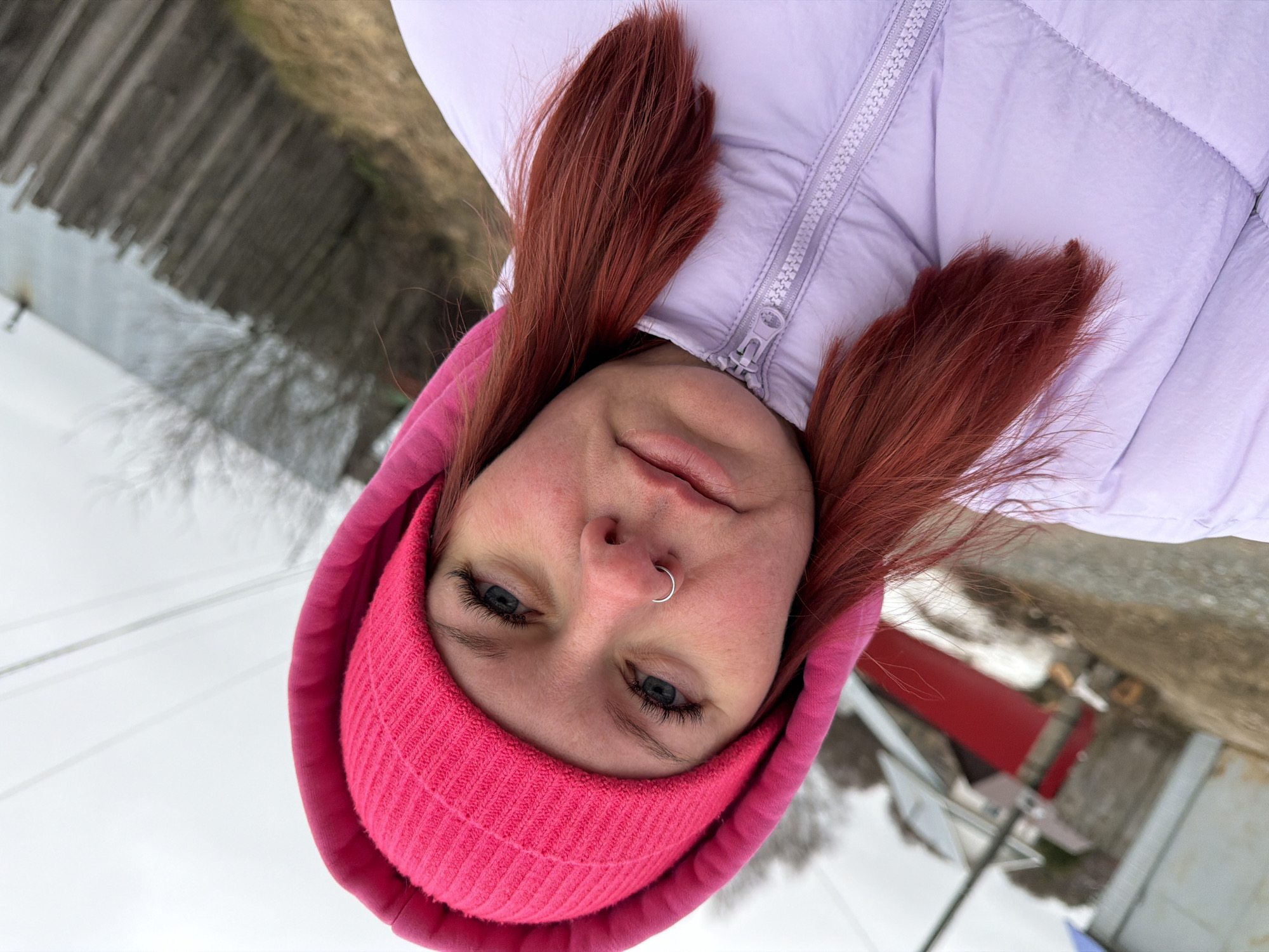 The image shows a person wearing a pink beanie and a light purple puffer jacket. The person has red hair and a silver nose ring. The background appears to be an outdoor environment with a blurred fence and possible building or structure with red paint visible. The location is Chaikovsky, Russia.