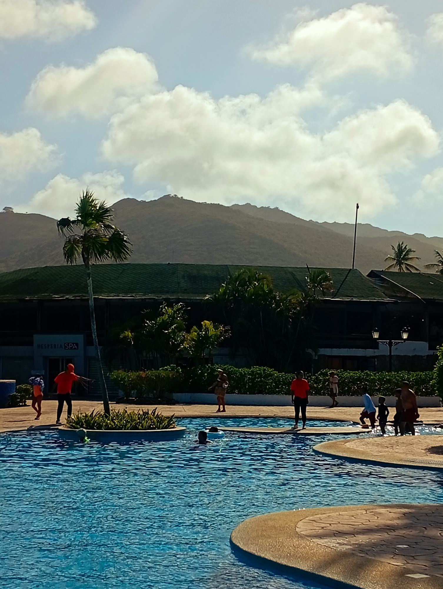 The image shows a swimming pool area in what appears to be a resort or hotel complex. The location is likely Playa El Agua, Venezuela, inferred from the "Hesperia Spa" signage visible on the building.
People of various ages are present, some near the pool's edge and others in the water. One individual is wearing an orange shirt. Several people are walking along the paved area surrounding the pool.
The environment includes lush green foliage, palm trees, and a mountain range in the background. The sky is partly cloudy. The pool's water is blue and appears clean. A structure with a dark green roof sits behind the pool area. There are lampposts present, indicating that the area is accessible during the evening or night.