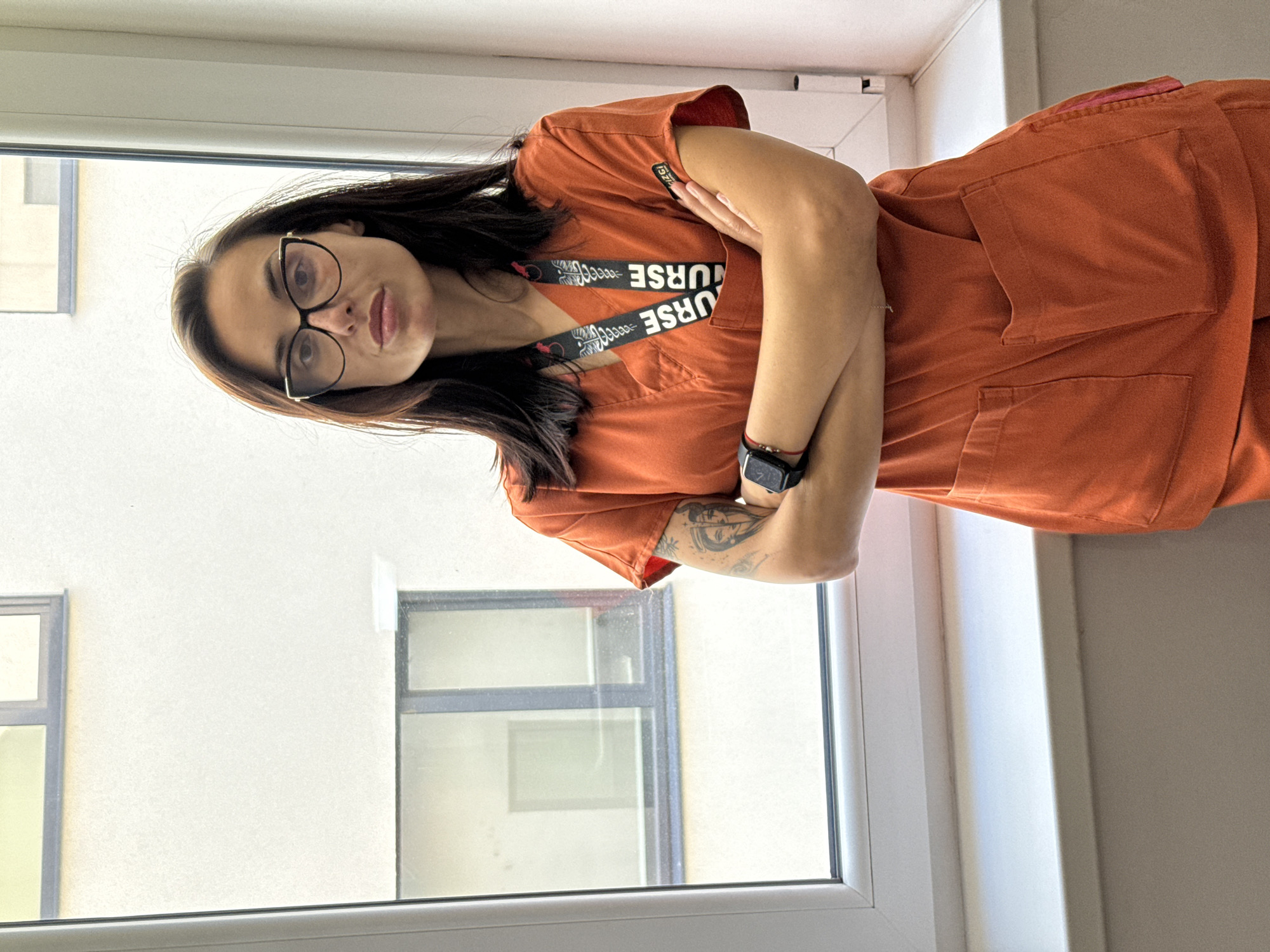 A woman with glasses stands indoors near a window. She is wearing an orange scrub top with a "NURSE" lanyard around her neck. She has a watch on her left wrist and a tattoo on her left forearm. The location is Pleven, Bulgaria, based on the fact the image was taken there. The woman is crossing her arms, and the window reveals part of a building across from it.