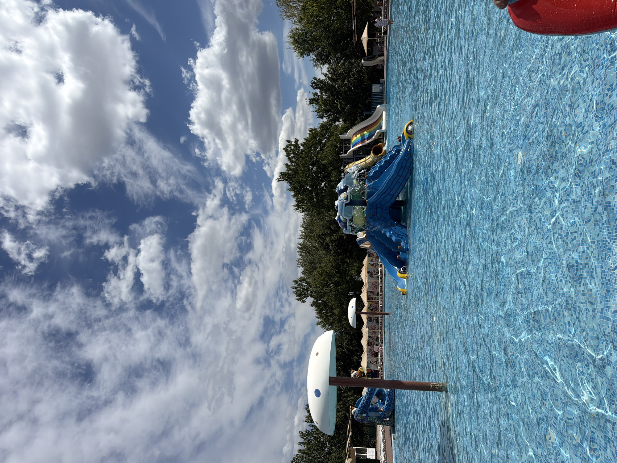 A wide shot of an outdoor swimming pool at a water park on a cloudy day. The pool is filled with clear blue water, and the bottom is tiled with small blue and white squares. In the center of the pool, there is a large blue and yellow inflatable water slide with multiple ramps. To the left of the slide, a blue inflatable serpent-like structure is partially submerged in the water. Two large, white mushroom-shaped umbrellas with blue dots are positioned in the pool. In the background, a line of beige lounge chairs and umbrellas are set up under a row of green trees. A few people are visible in the distance, relaxing near the lounge chairs. The sky is a bright blue with scattered white, fluffy clouds. The water park appears to be located in Sociteni, Moldova.