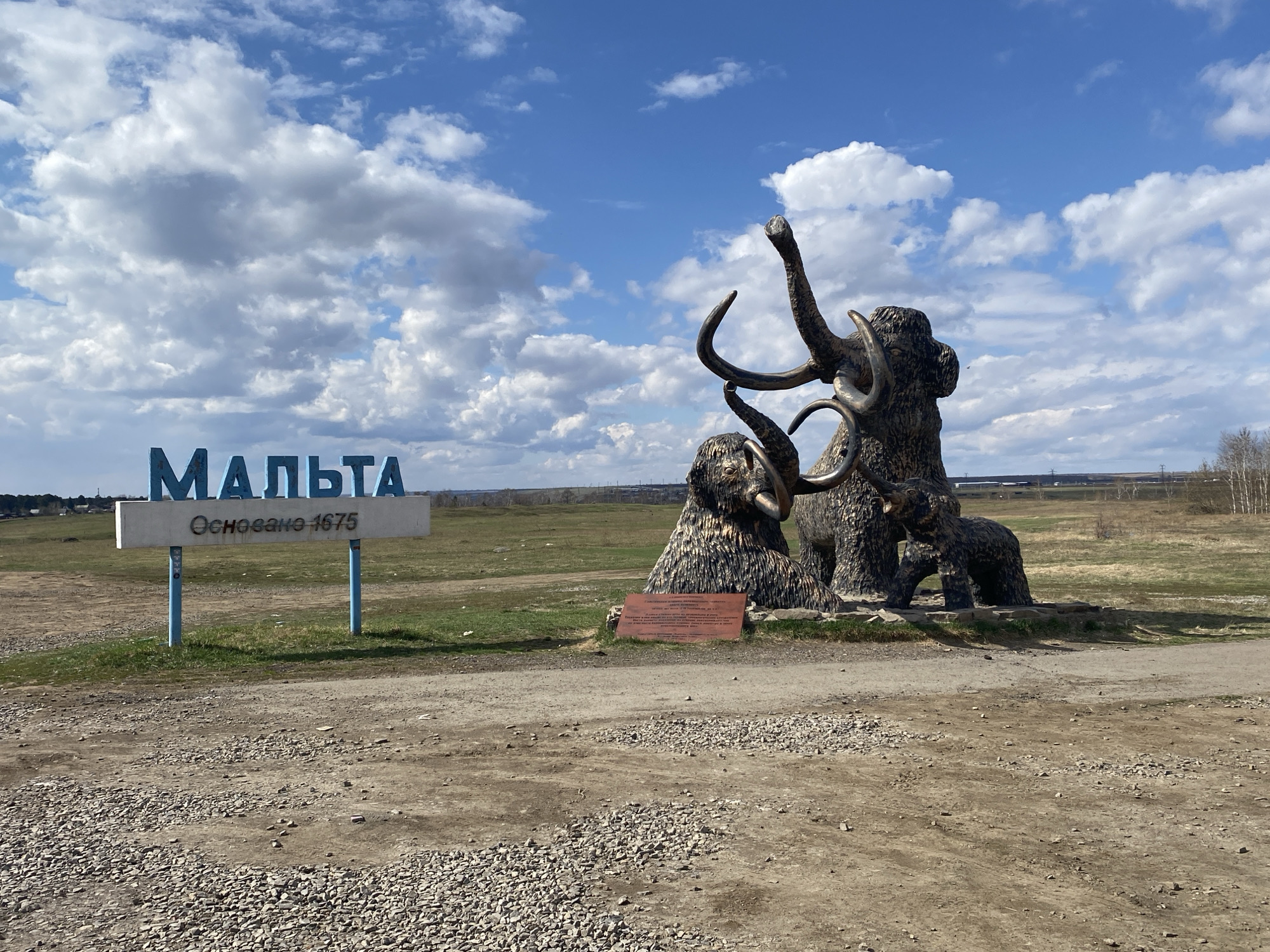 The image depicts a roadside scene in Malta, Russia. A sign in Russian, "Мальта" (Malta) and "Основано 1675" (Founded 1675), marks the entrance to the town. To the right of the sign is a sculpture of a family of mammoths, including an adult and two smaller mammoths. The ground is gravel and dirt, with fields and sparse trees in the background. The sky is blue with scattered white clouds.