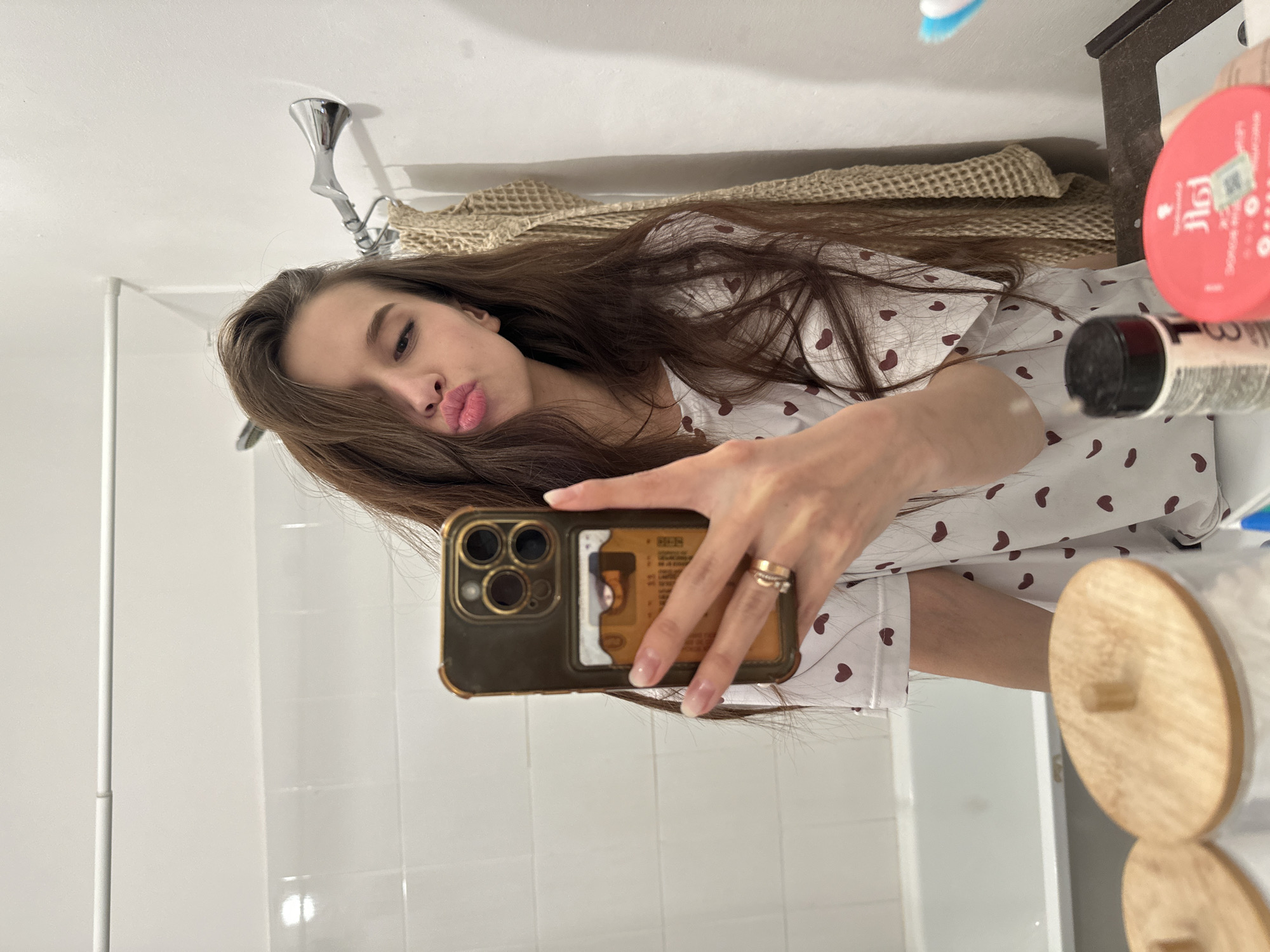 A woman is taking a selfie in a bathroom. She is wearing a white pajama top with small heart patterns. She is pouting for the camera while holding a gold-colored smartphone. The bathroom has white tiled walls and a washbasin with toiletries on it. A decorative wall hanging is visible in the background. The location, city, or country cannot be verified from this image.
