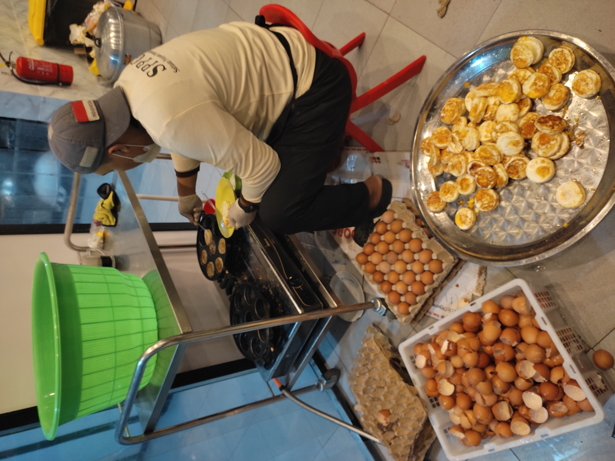 In Ponorogo, Indonesia, a person is cooking small, round, fried items, likely quail eggs, in a specialized pan on a portable stovetop. The setup appears to be outdoors or in an open-air market area, indicated by the tiled floor and a glimpse of a building entrance or window. A large green plastic basin sits on a stainless steel cart, possibly for washing or holding ingredients. Nearby, trays of whole eggs and a white plastic basket filled with eggshells are visible, suggesting preparation is underway. The cook, wearing a white long-sleeved shirt with a logo, a gray baseball cap with an Indonesian flag patch, and gloves, is actively engaged in the cooking process, using a yellow plate and tongs. A bright red child's chair is positioned next to the cook. In the background, a red fire extinguisher is mounted on a wall.