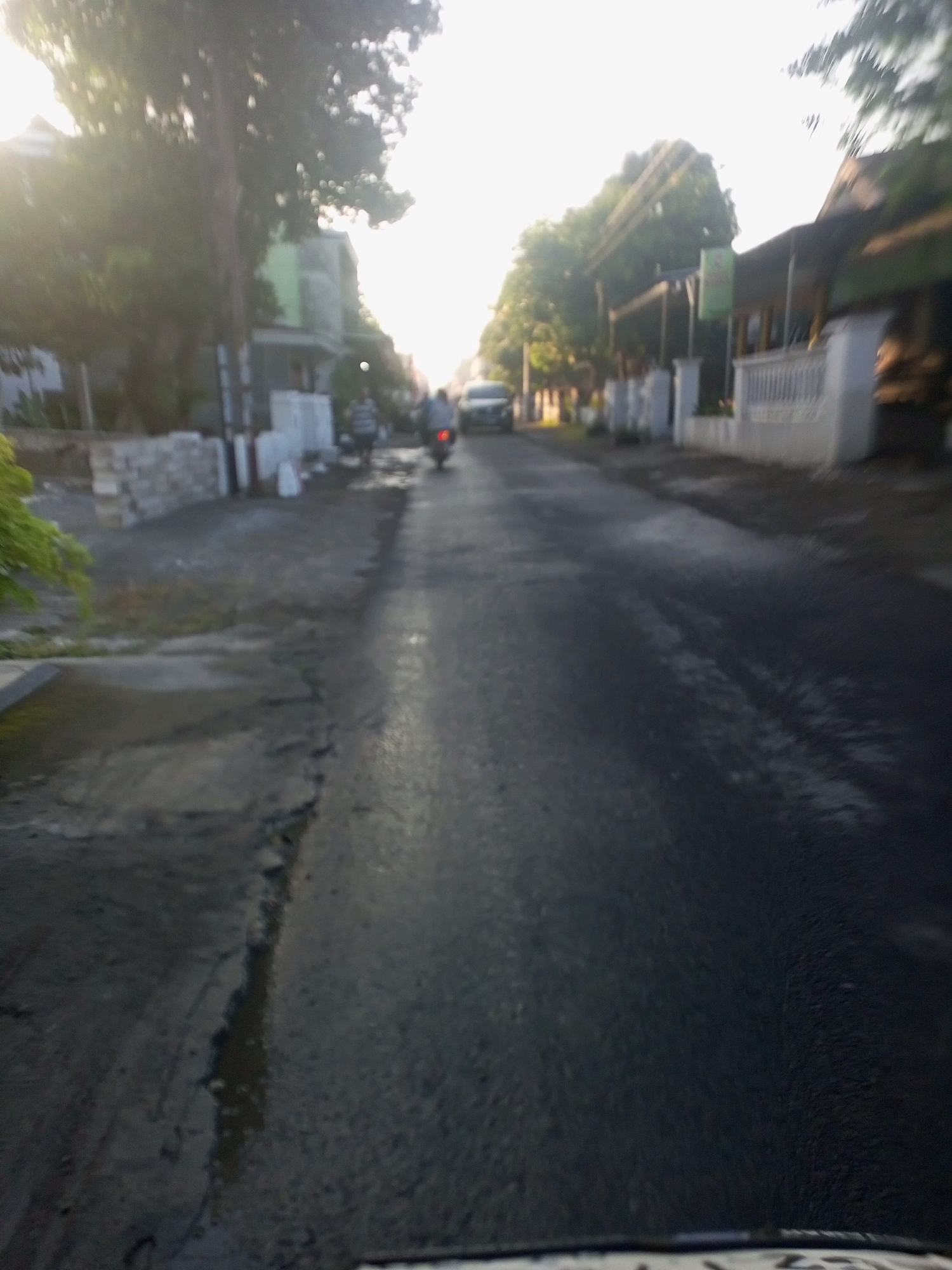 The image captures a street scene in Ngasem, Indonesia, characterized by a slight blur and a bright, hazy sky, suggesting it might be early morning or late afternoon. The road surface appears wet and reflective, with puddles and what looks like cracks or uneven patches. On the left side of the road, there are buildings and some vegetation, including what seems to be a leafy plant in the foreground. White posts or pillars line the side of the road in several places, and some low walls are visible.
In the middle ground, two motorcycles are present. One is ridden by a person wearing a dark shirt and light-colored pants, and the other is ridden by a person in a light-colored shirt and dark pants. Both individuals are moving away from the camera. Further down the road, a car is visible, also in motion.
The overall impression is of a quiet, possibly residential or commercial street in Ngasem, Indonesia, during a period of moisture on the road.