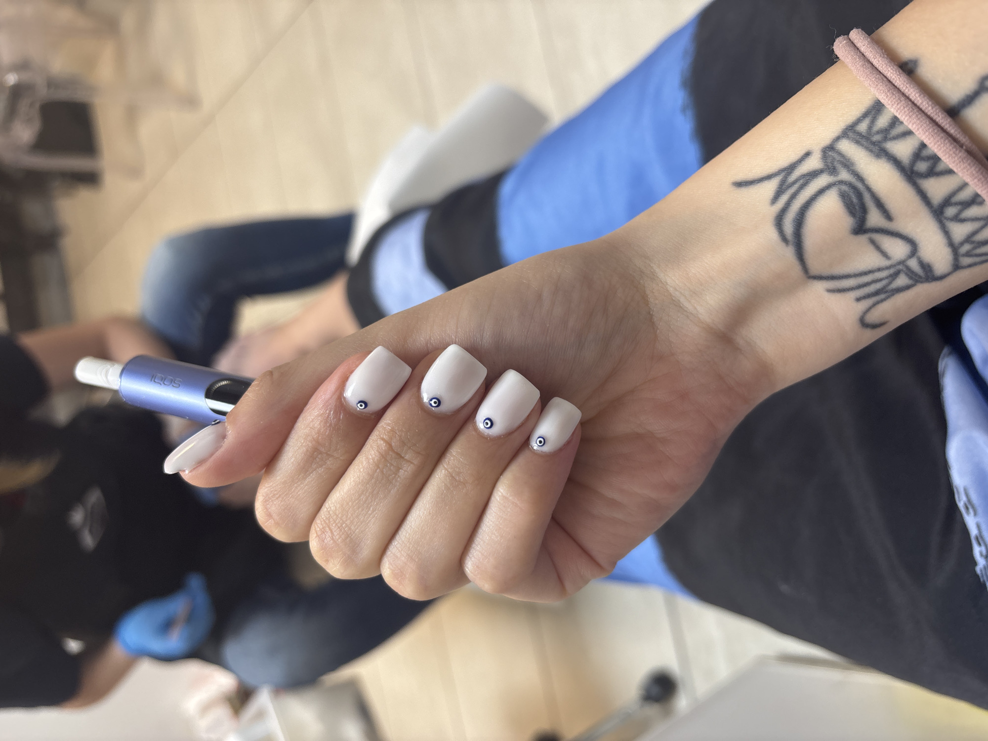 A person is seated, with their left arm visible in the foreground. The arm has a tattoo near the wrist depicting a heart, crown and other indiscernible components, and is adorned with a pink hair tie. The person's fingernails are painted white with small blue "evil eye" designs on the middle three fingers. The person is holding an IQOS heated tobacco product.
The background is blurry, suggesting the image was taken indoors. The environment includes elements of a salon or spa setting; a person wearing blue gloves can be partially seen on the left. Given the stated location, the image was taken in Sofia, Bulgaria.