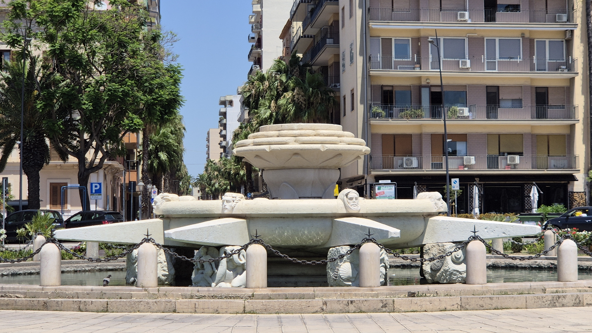 The image depicts a stone fountain located in Taranto, Italy, likely in a public square. The fountain is the central focus, with intricate carvings of faces and figures adorning its structure. It appears to be made of light-colored stone and is surrounded by water. In the background, there are multi-story buildings with balconies and windows, typical of urban architecture. Palm trees and other greenery are visible, suggesting a warm climate. Several cars are parked along the street in front of the buildings. A pigeon is present near the fountain, adding a touch of life to the scene. The overall atmosphere suggests a sunny day in a Mediterranean city.