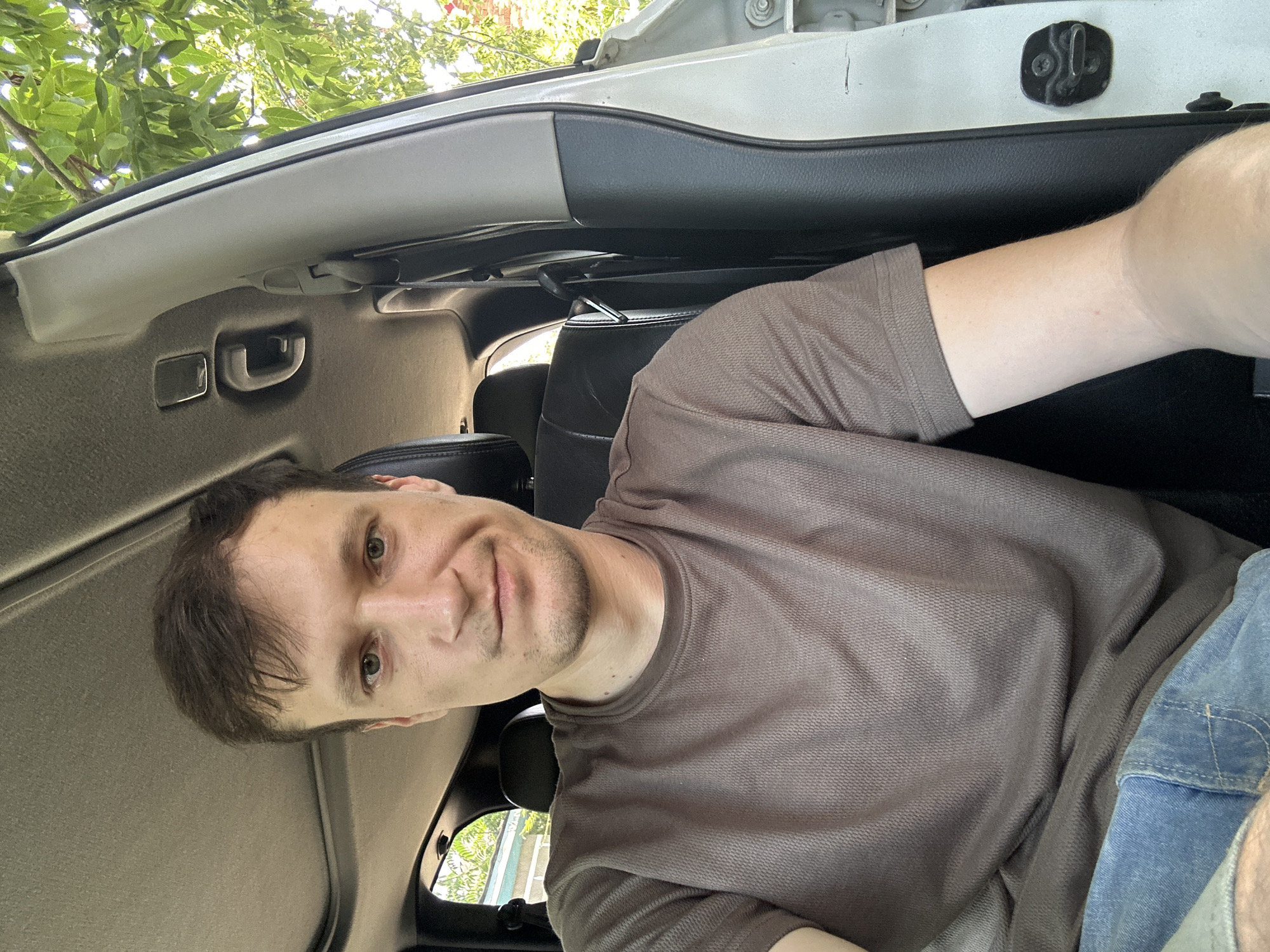 A man is taking a selfie inside a vehicle. He is wearing a brown t-shirt and blue jeans. The man is looking directly at the camera with a neutral expression. The interior of the vehicle is visible, including the seats, ceiling, and door frame. Outside the vehicle, green foliage and trees can be seen. The image appears to have been taken during the daytime. There is no information in the image to indicate the location in the city of Makiv, Ukraine.