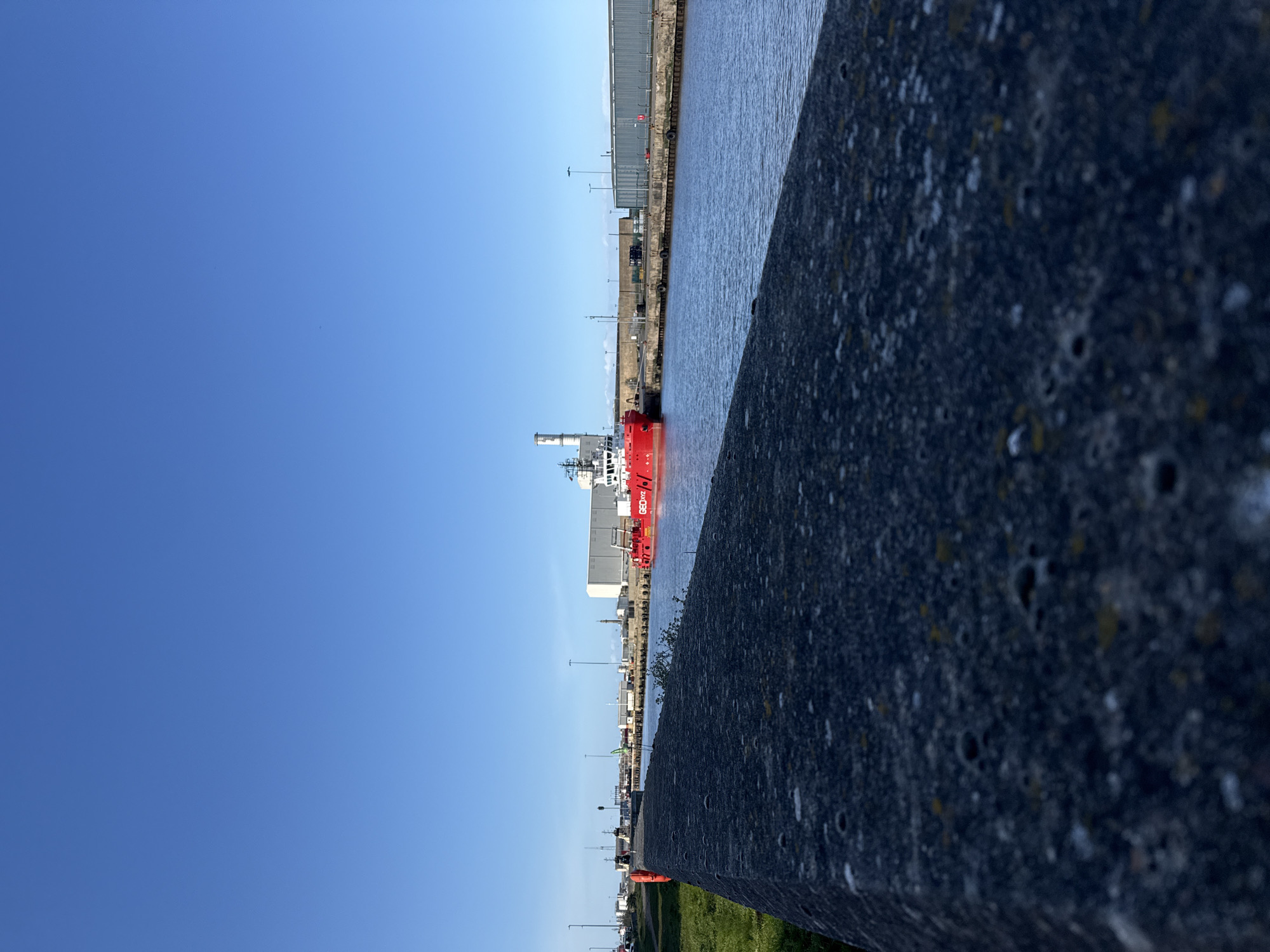 This outdoor shot depicts a maritime scene in Gorleston-on-Sea, UK. A red and white ship is docked in the water, viewed from an elevated perspective. A textured, dark grey stone wall dominates the foreground, partially obscuring the view. Behind the ship, buildings and structures line the waterfront. Streetlights and vehicles are visible along a road running parallel to the water. Above, a clear blue sky fills the remaining space.