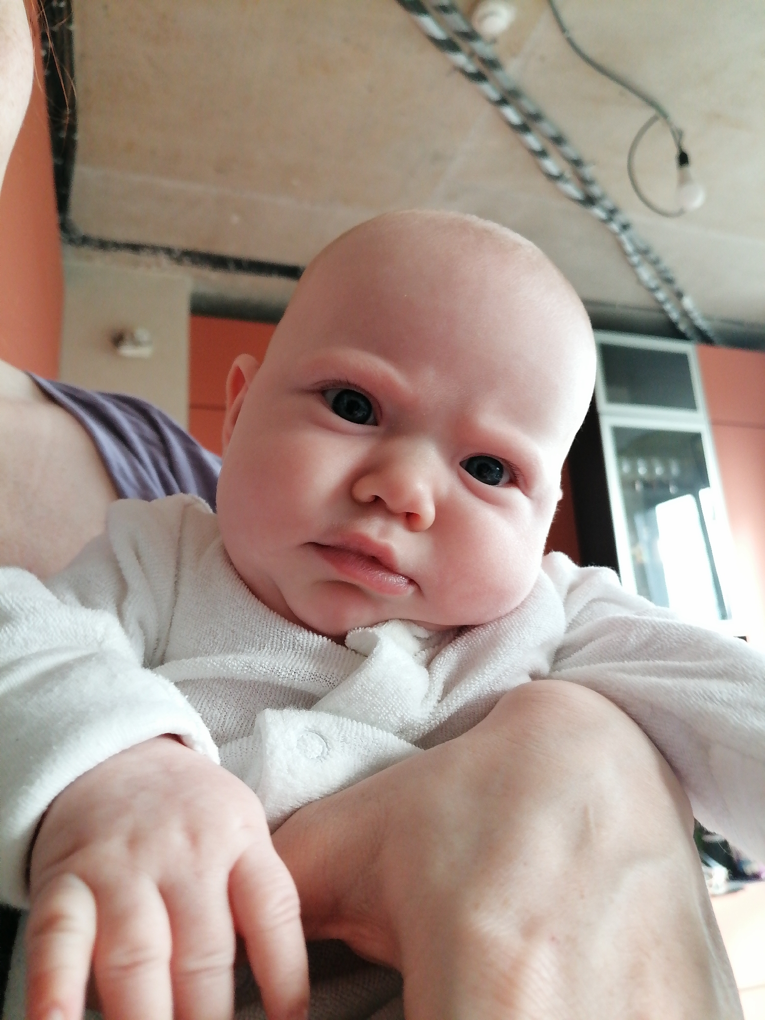 The image shows a baby being held. The baby has a white complexion, a bald head, and is wearing a white, long-sleeved garment. The baby has a serious or contemplative expression. The baby is being held by a person, and their arm and hand are visible. The person's arm has a light skin tone. The person is wearing a lilac-colored top.
The background includes a partially visible wall that is painted with some orange tones, and a white surface with exposed wiring and light fixtures. A partially visible door or window frame is in the background as well.
No specific location in any city or region is discernible from this image.
