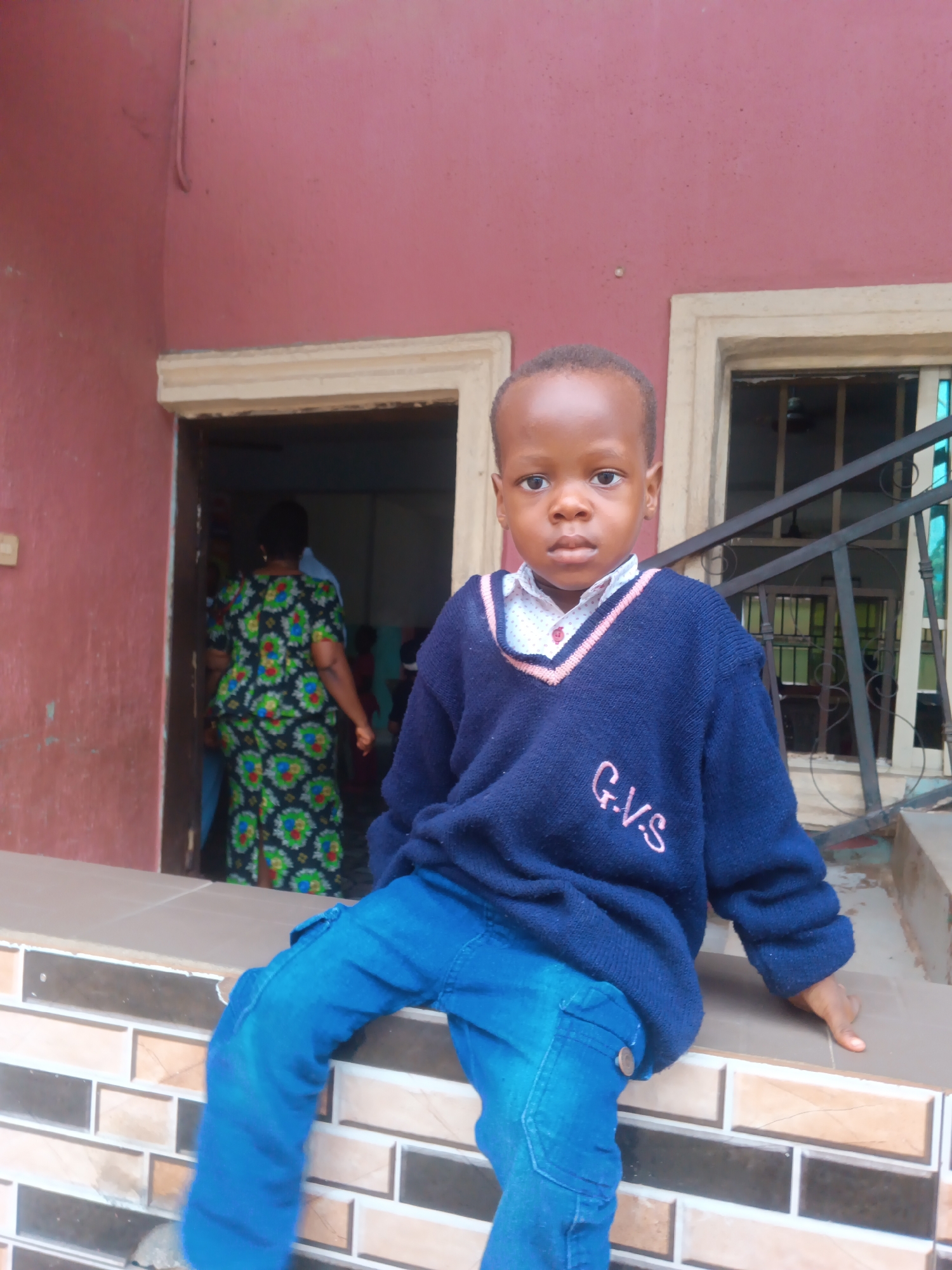 A young boy, appearing to be of African descent, is seated on a tiled ledge in front of a building. He is wearing a dark blue V-neck sweater over a white collared shirt with a subtle pattern, and blue jeans. The sweater has pink lettering on the chest that reads "G.V.S." Behind him, an open doorway reveals a woman in a vibrantly patterned dress and other indistinct figures inside. The building exterior is painted a reddish-pink color, and a window with a metal railing is visible to the right. The tiled ledge appears to be part of a veranda or entryway. The overall setting suggests an outdoor urban environment, possibly in Nkpor, Nigeria, given the architectural style and common attire.